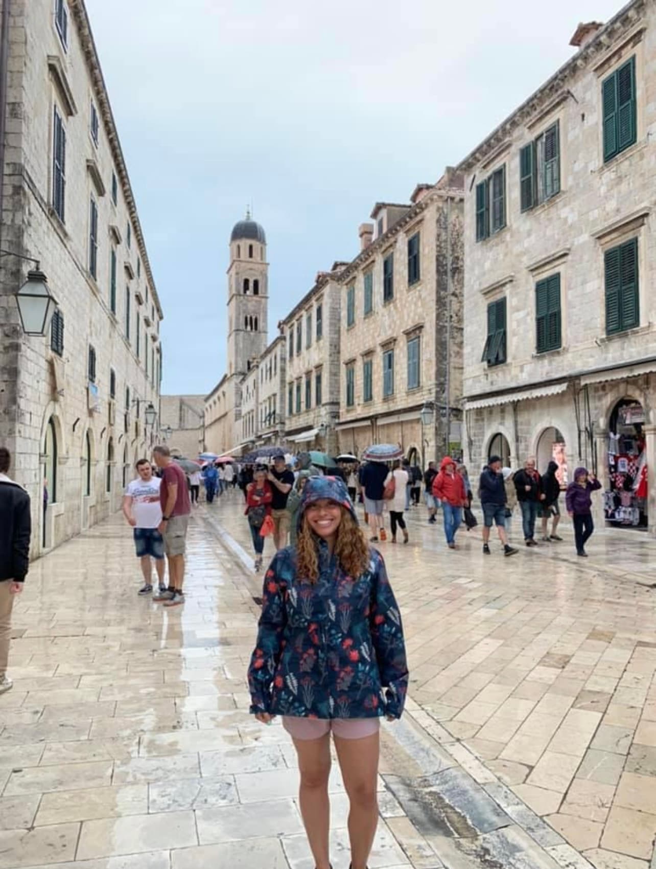 Maria in a raincoat posing on a street lined with white buildings with other pedestrians visible behind her