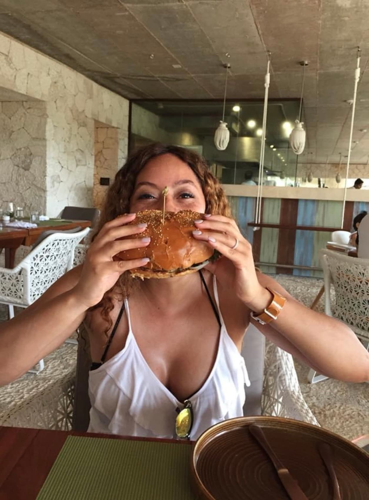 Maria in a white top sitting at a table holding a large sandwich in front of her face