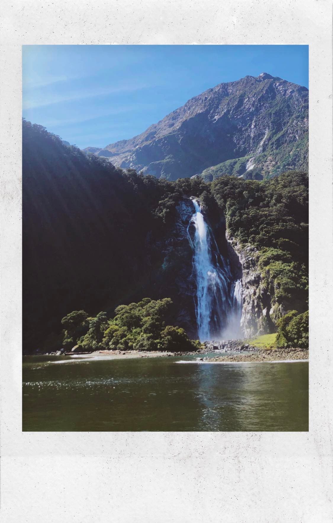 Polaroid photo of a far away view of a waterfall and mountains in the background during the daytime.