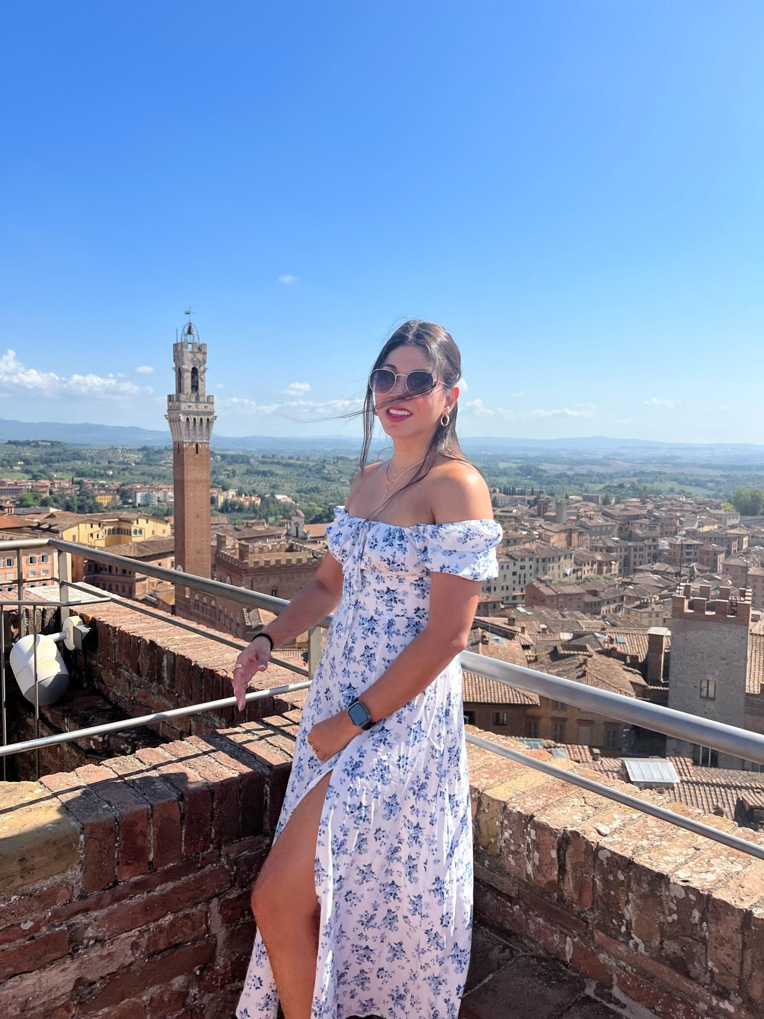 Carmen in a long white dress on a rooftop in Volterra on a sunny day with the cityscape visible behind her
