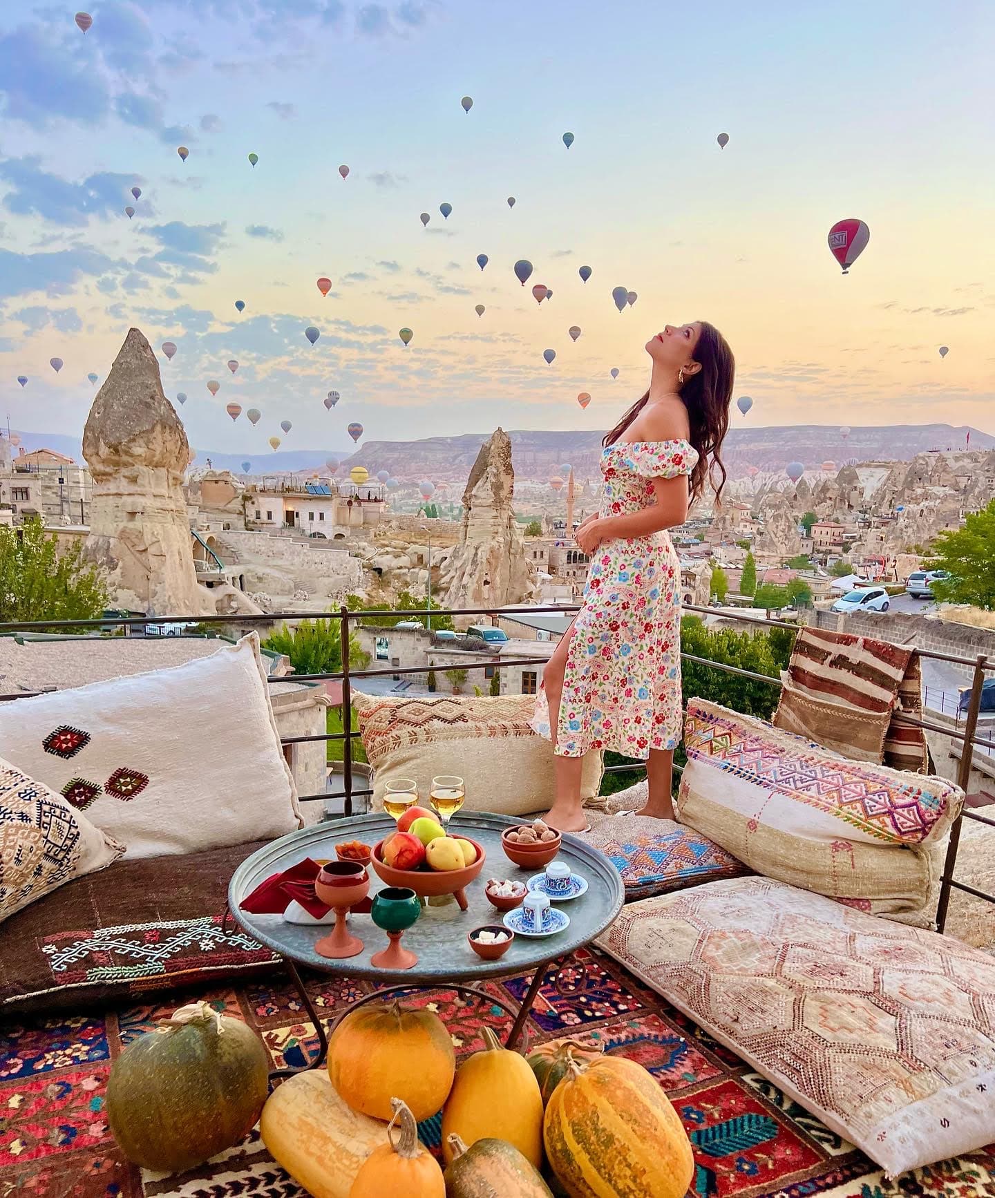 Carmen in a floral dress standing on a rooftop with the hot air balloons of Cappadocia floating in the distance