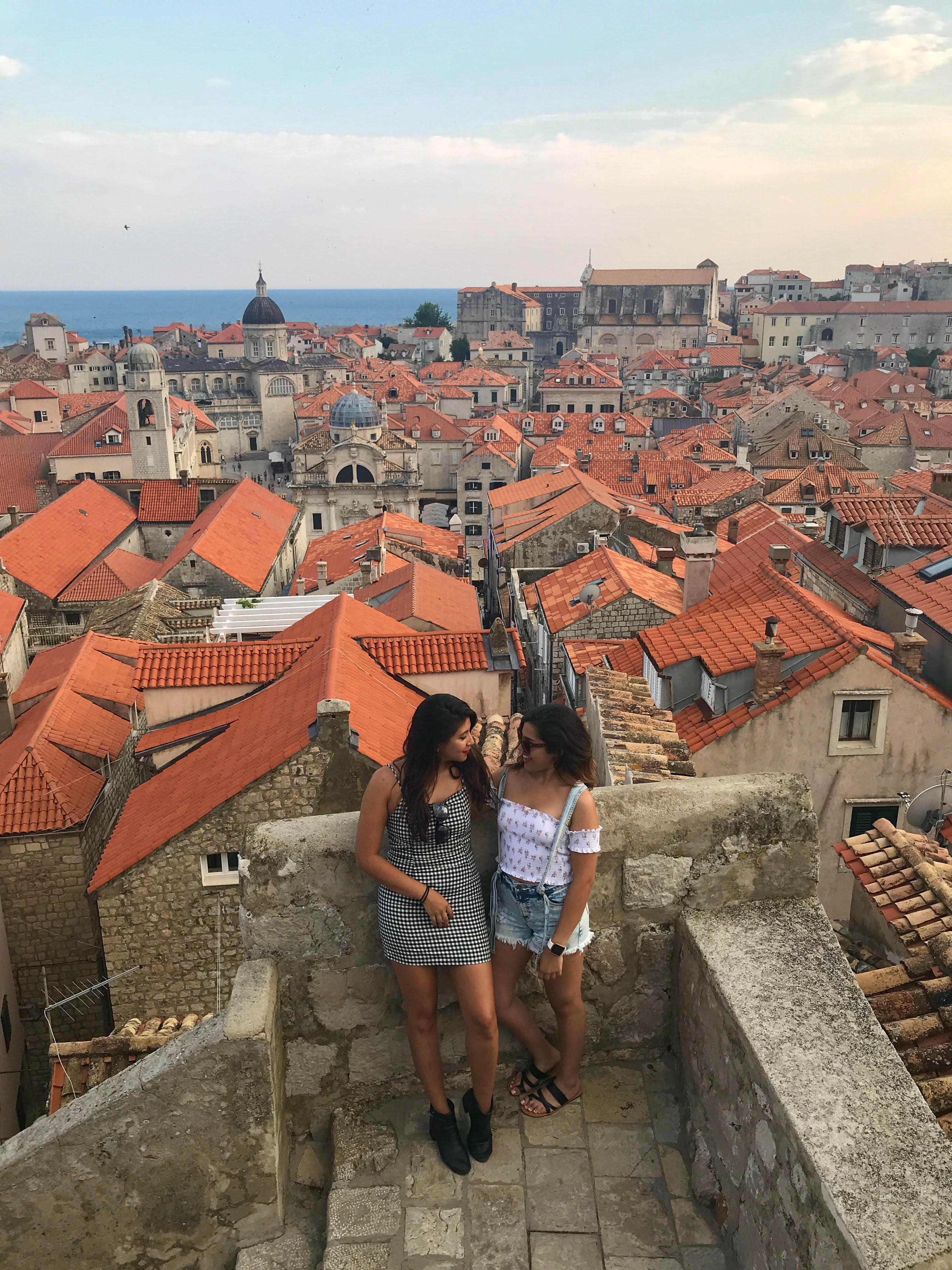 Carmen and a friend standing on a rooftop in Dubrovnik with many orange rooftops visible and the sea behind them