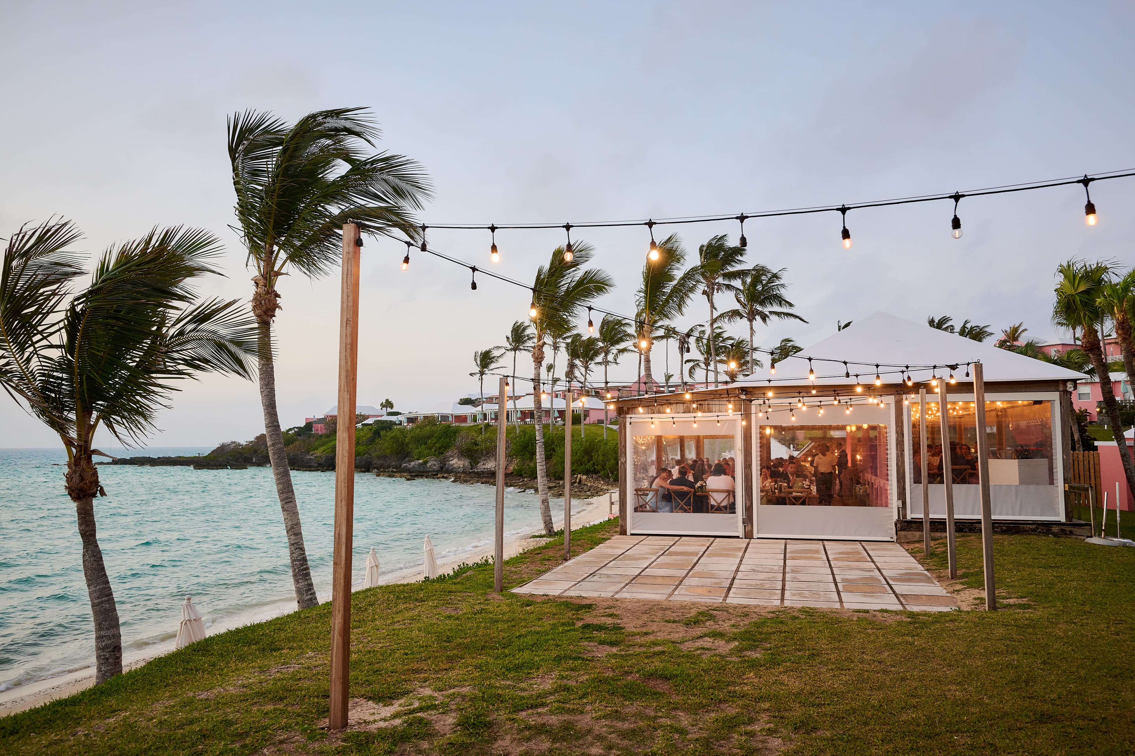 Pretty view of an outdoor patio with string lights hanging above right next to the sea and several palm trees