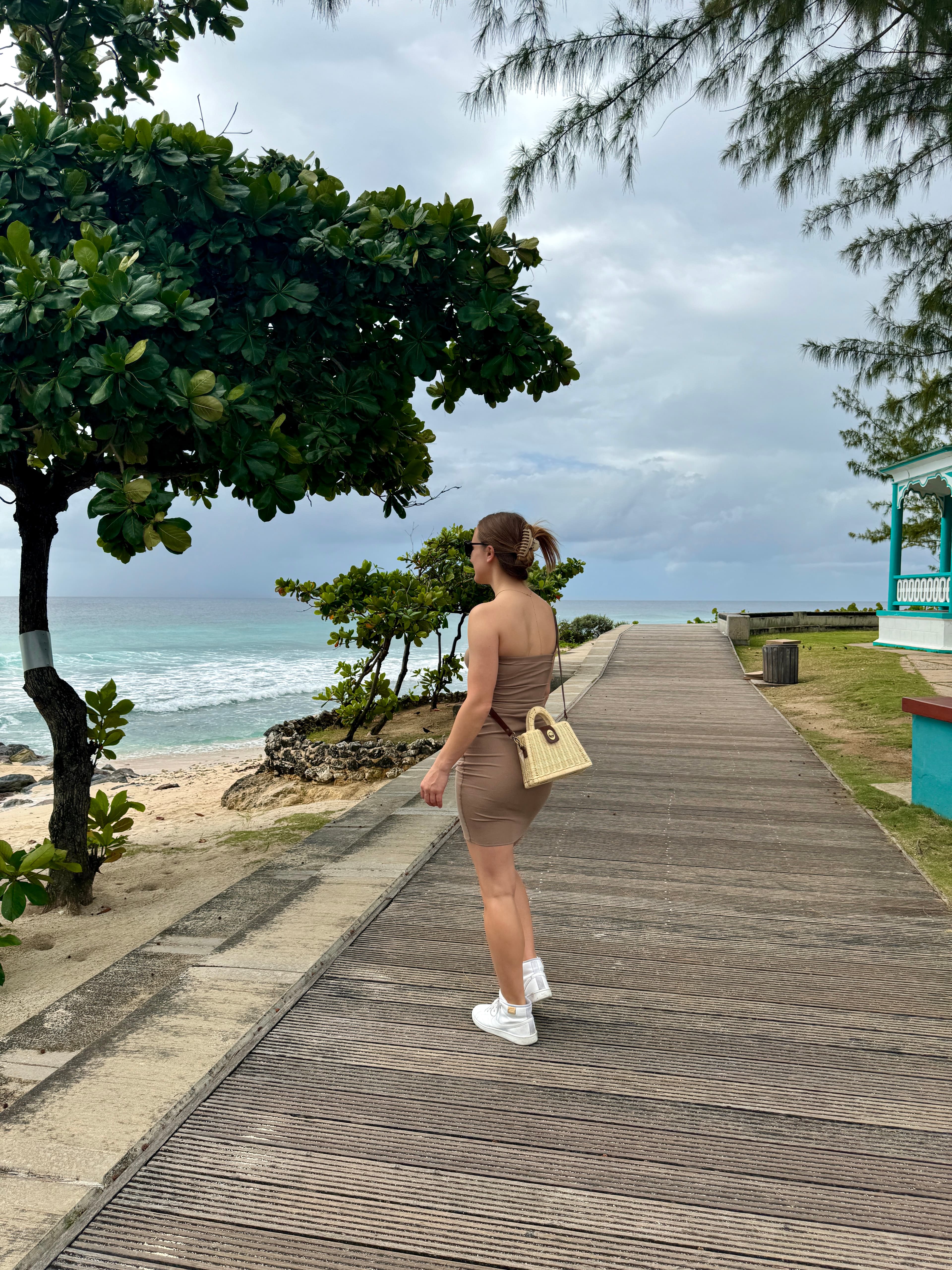 A woman posing on a boardwalk while she looks out to the sea and green trees.