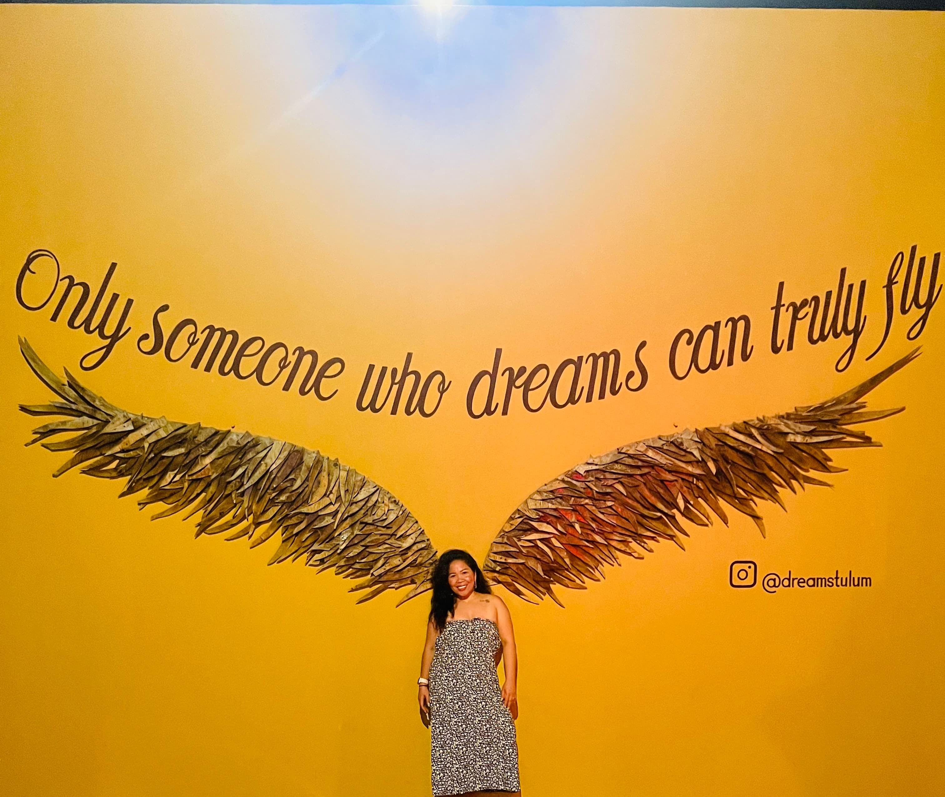 Rogelyn in a long dress posing against a wall mural that makes it look as if she has wings, reading “only someone who dreams can truly fly”