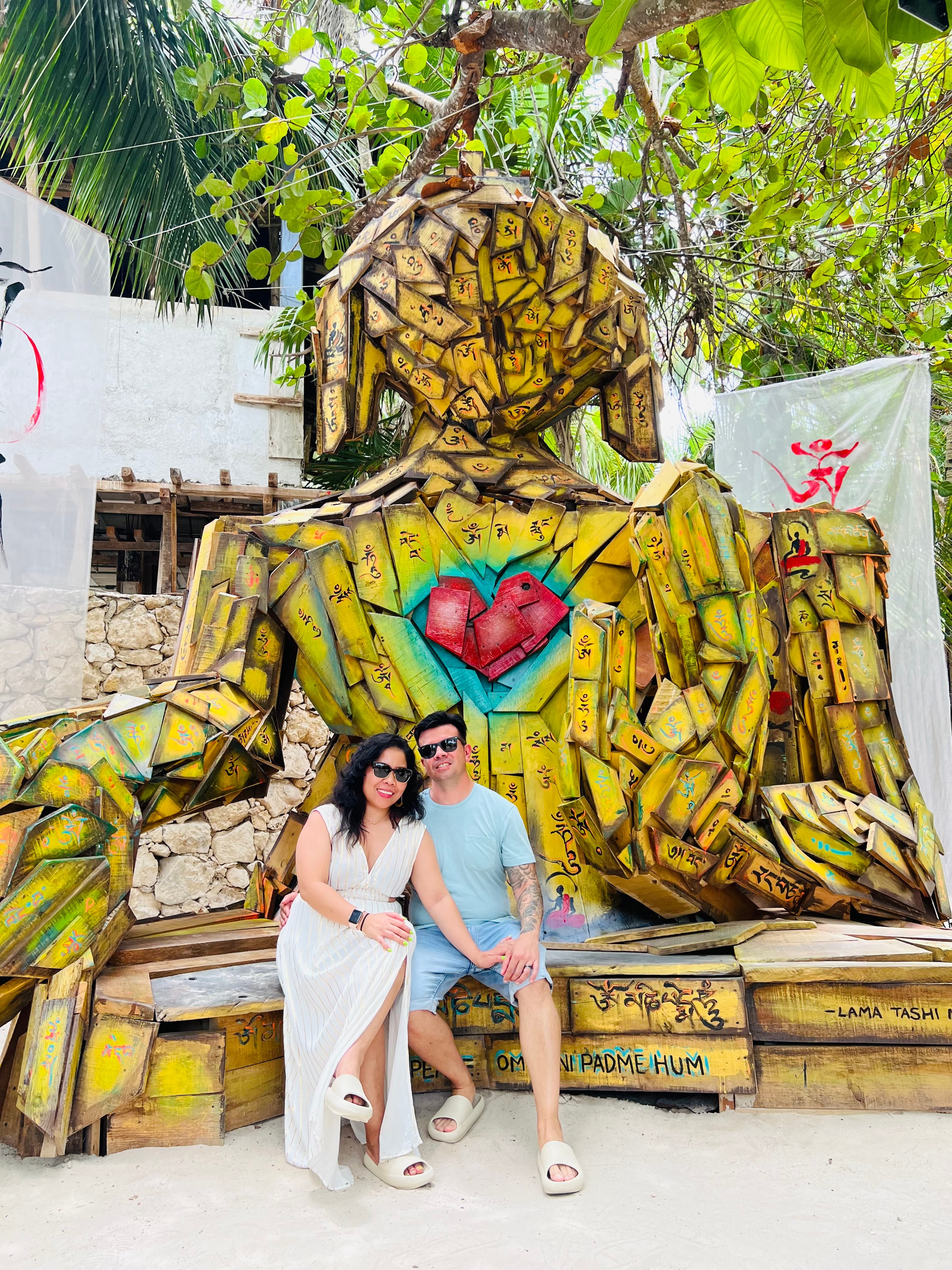 Rogelyn and a man posing for a photo in front of a yellow wooden sculpture shaped like a human