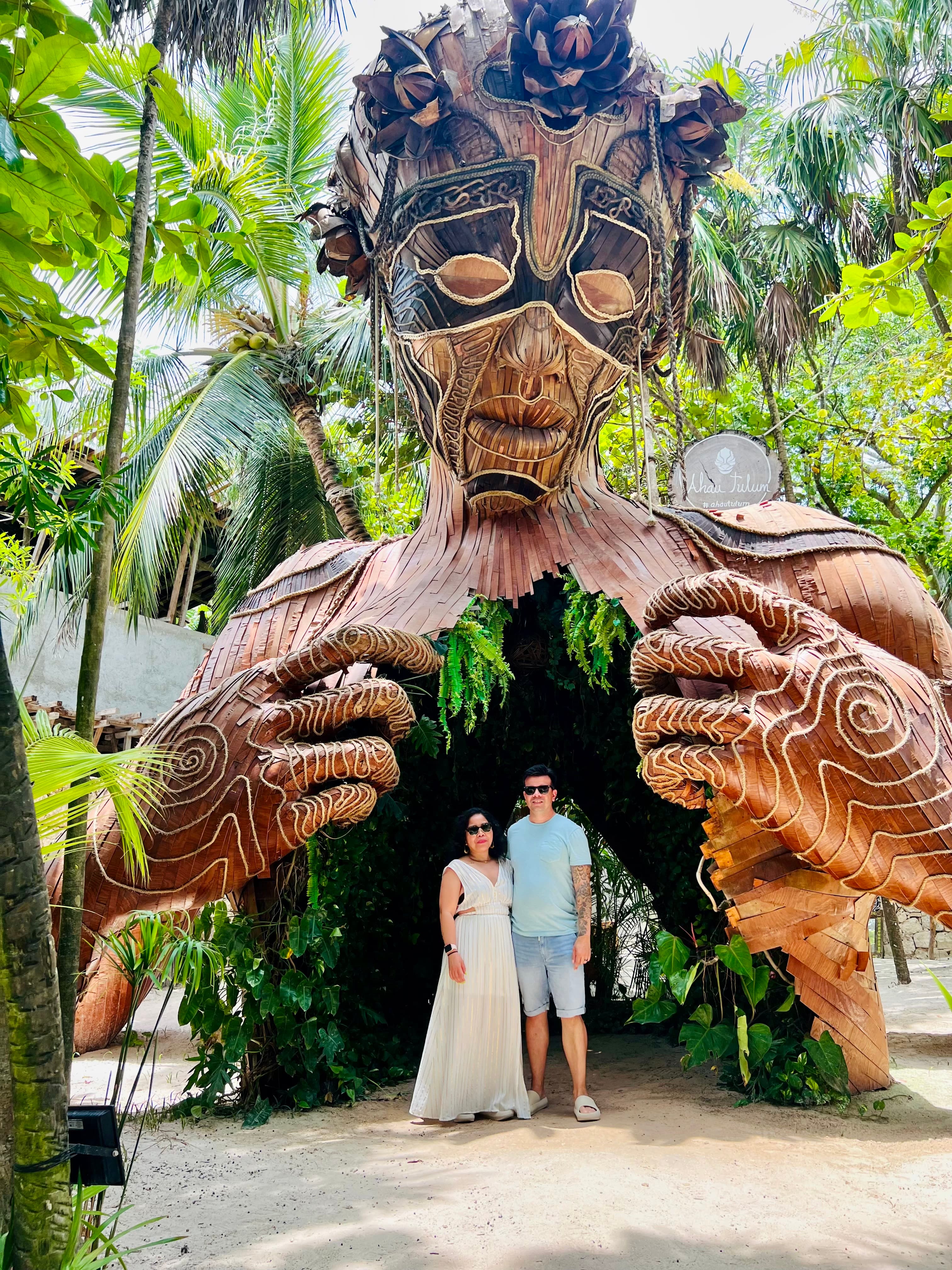 Rogelyn and a man with their arms around one another under a large wooden sculpture shaped like a human body