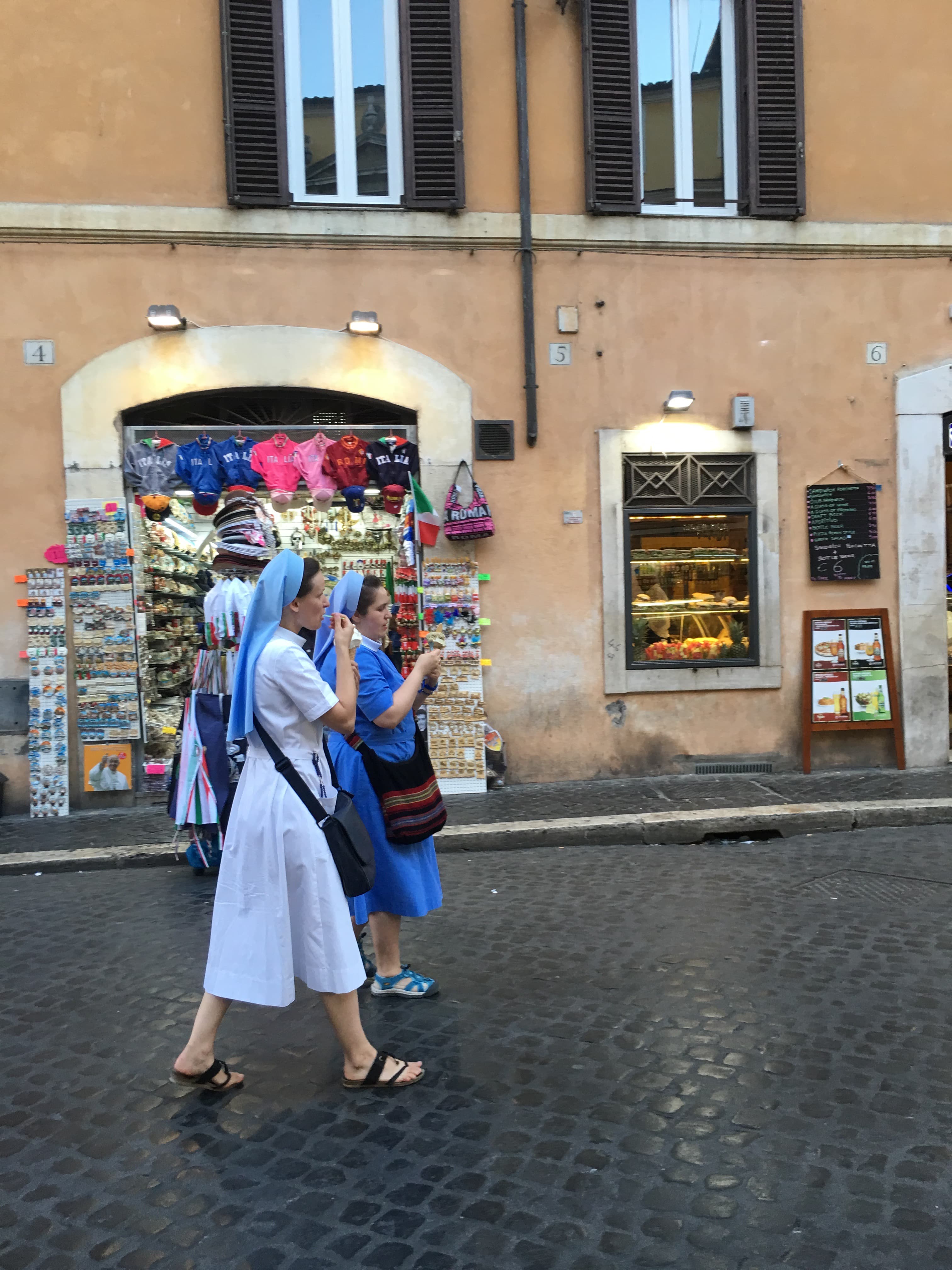 View of two women in blue headscarves walking side by side on a street in front of a beige building
