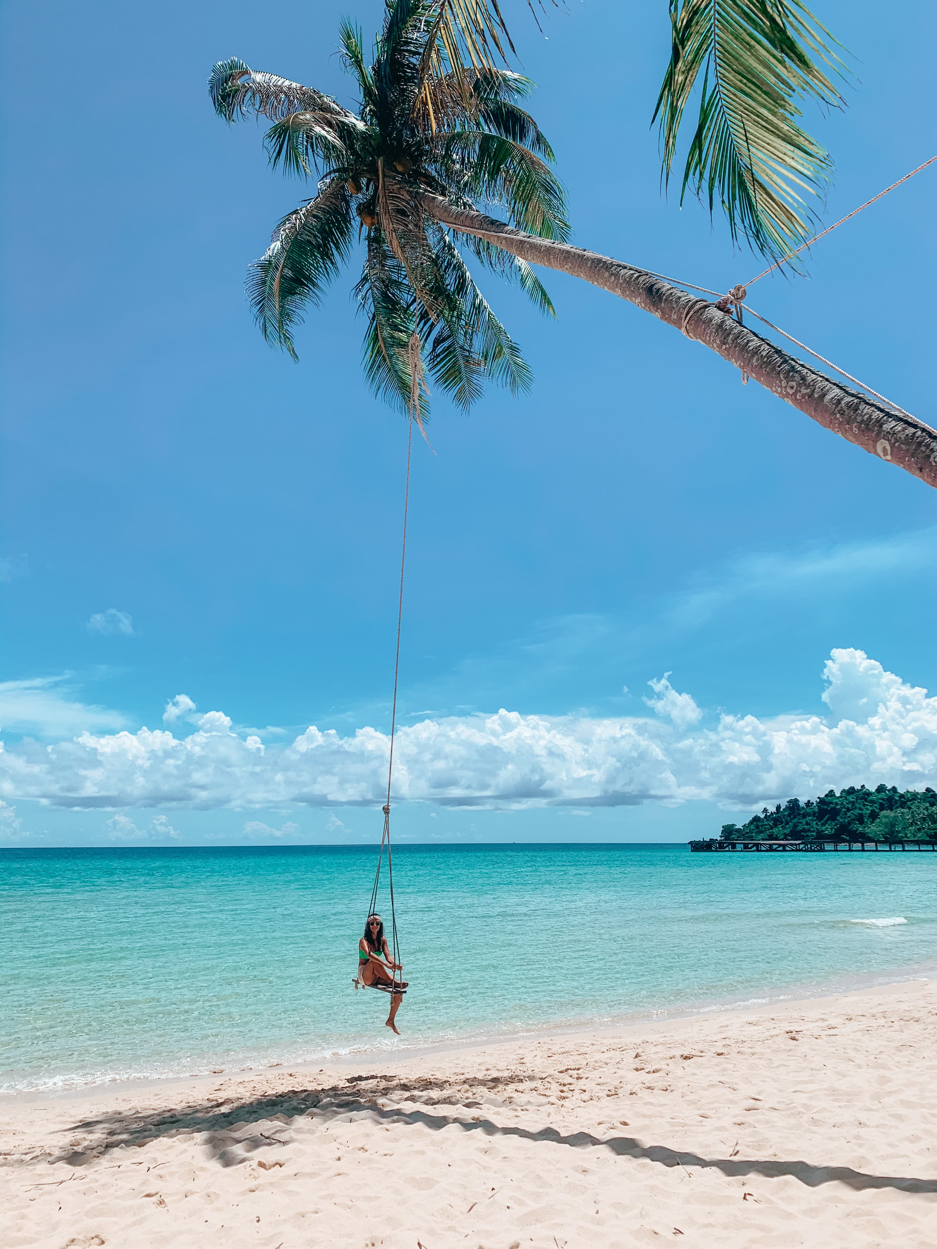 Janine sitting on a rope swing hanging from a slanted palm tree on a beautiful white sand beach
