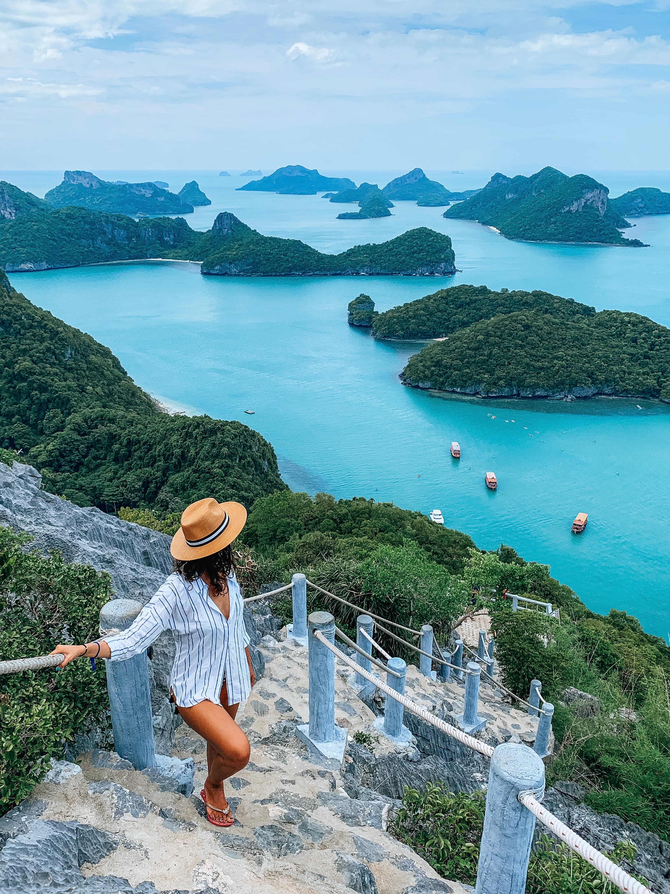 Janine in a white shirt and hat posing on stone steps leading down to a bright blue sea and surrounding islands