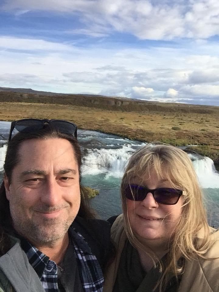 Kelli and a man taking a selfie outdoors in front of glaciers and a grassy field on a sunny day