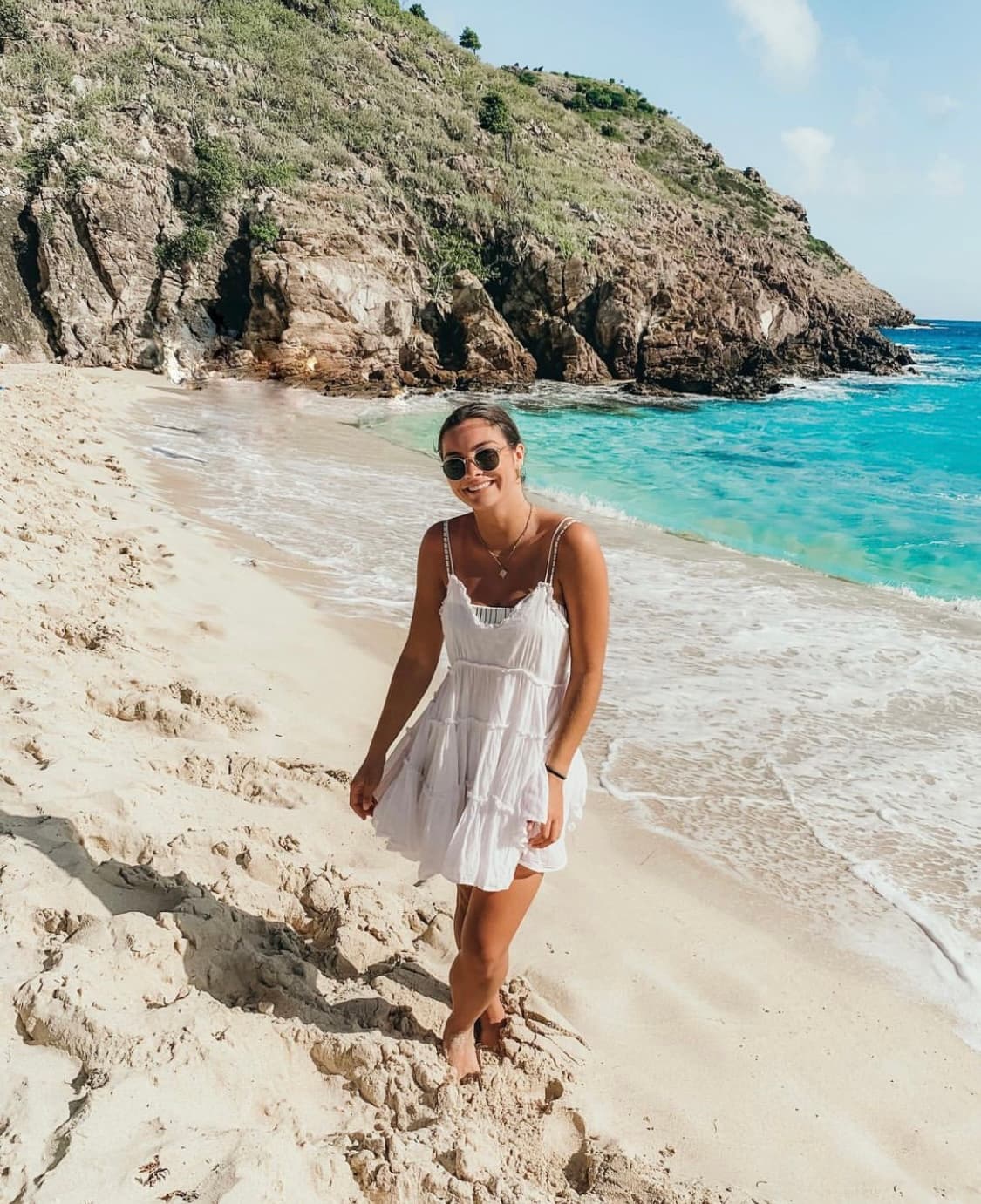 Advisor wearing a white dress on a beach with water and rocky cliffs in the background.