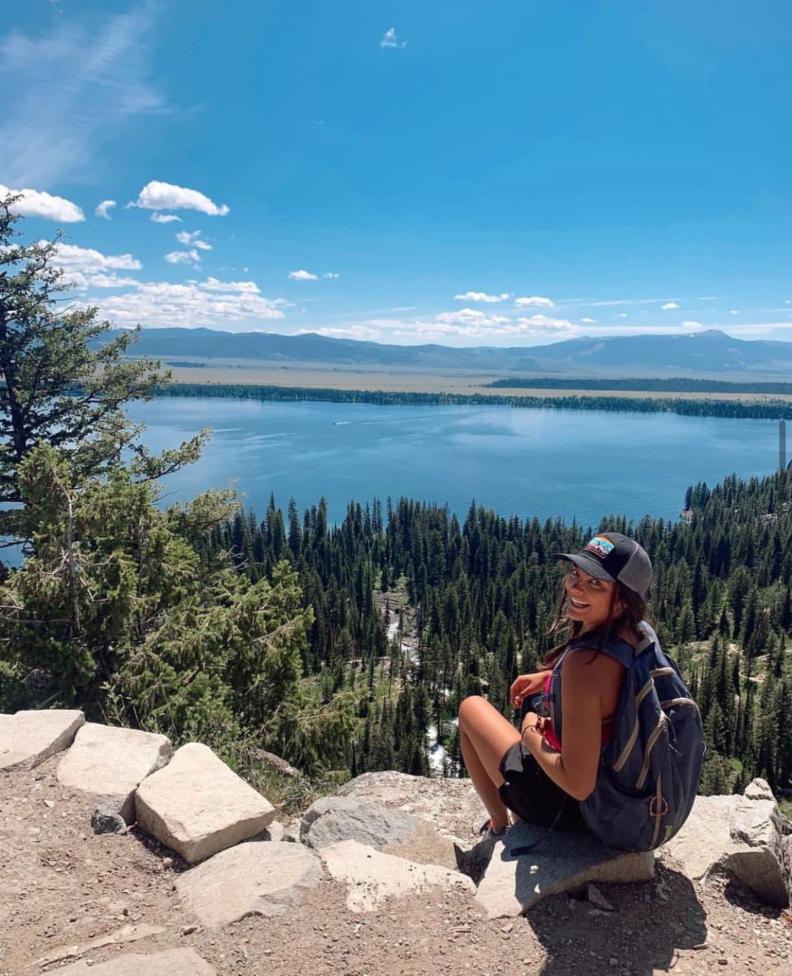 Advisor wearing a backpack on a ridge overlooking pine trees and a lake.