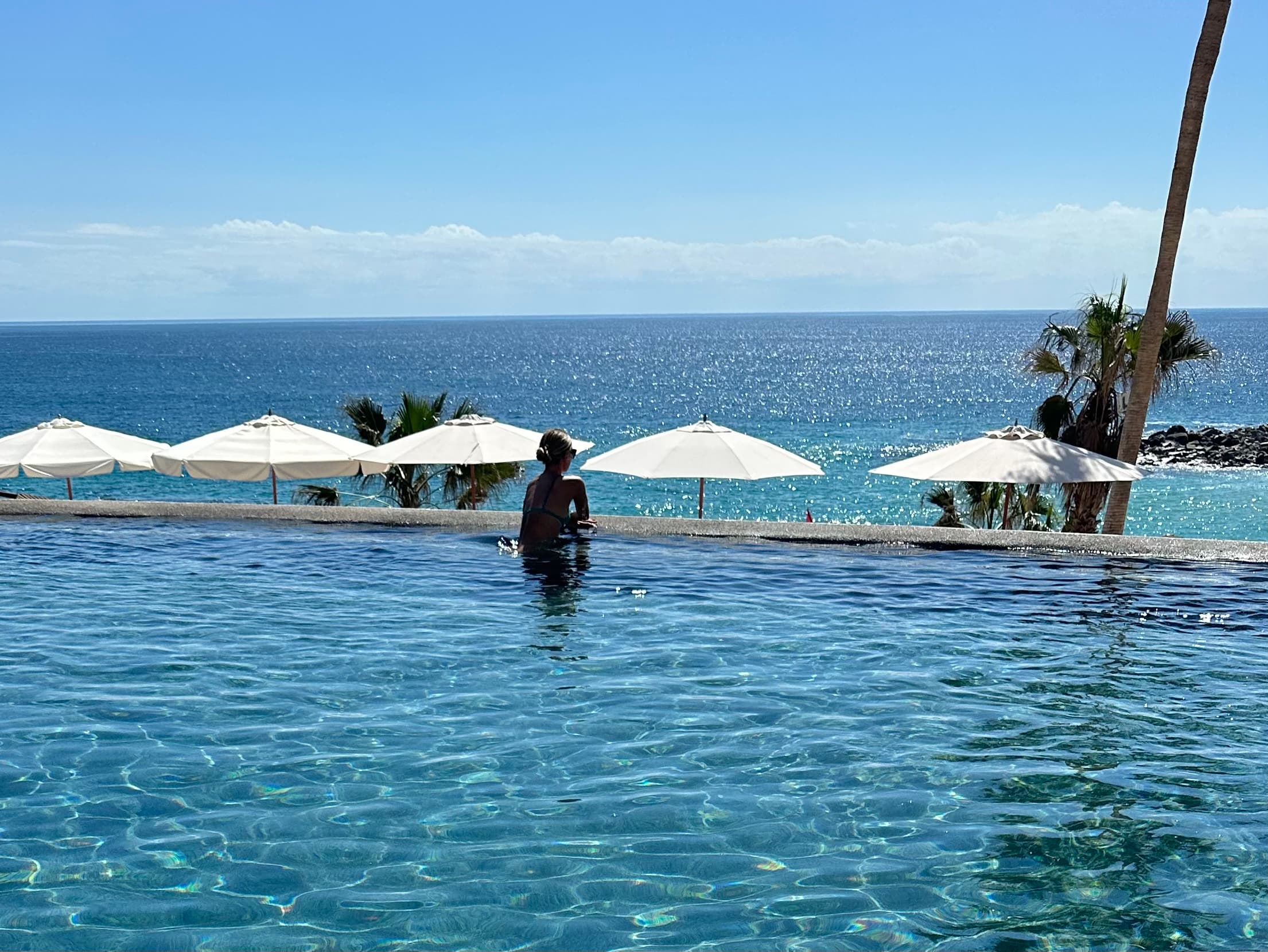 Advisor relaxing in an infinity pool with ocean, palm trees and sun umbrellas on a sunny day.