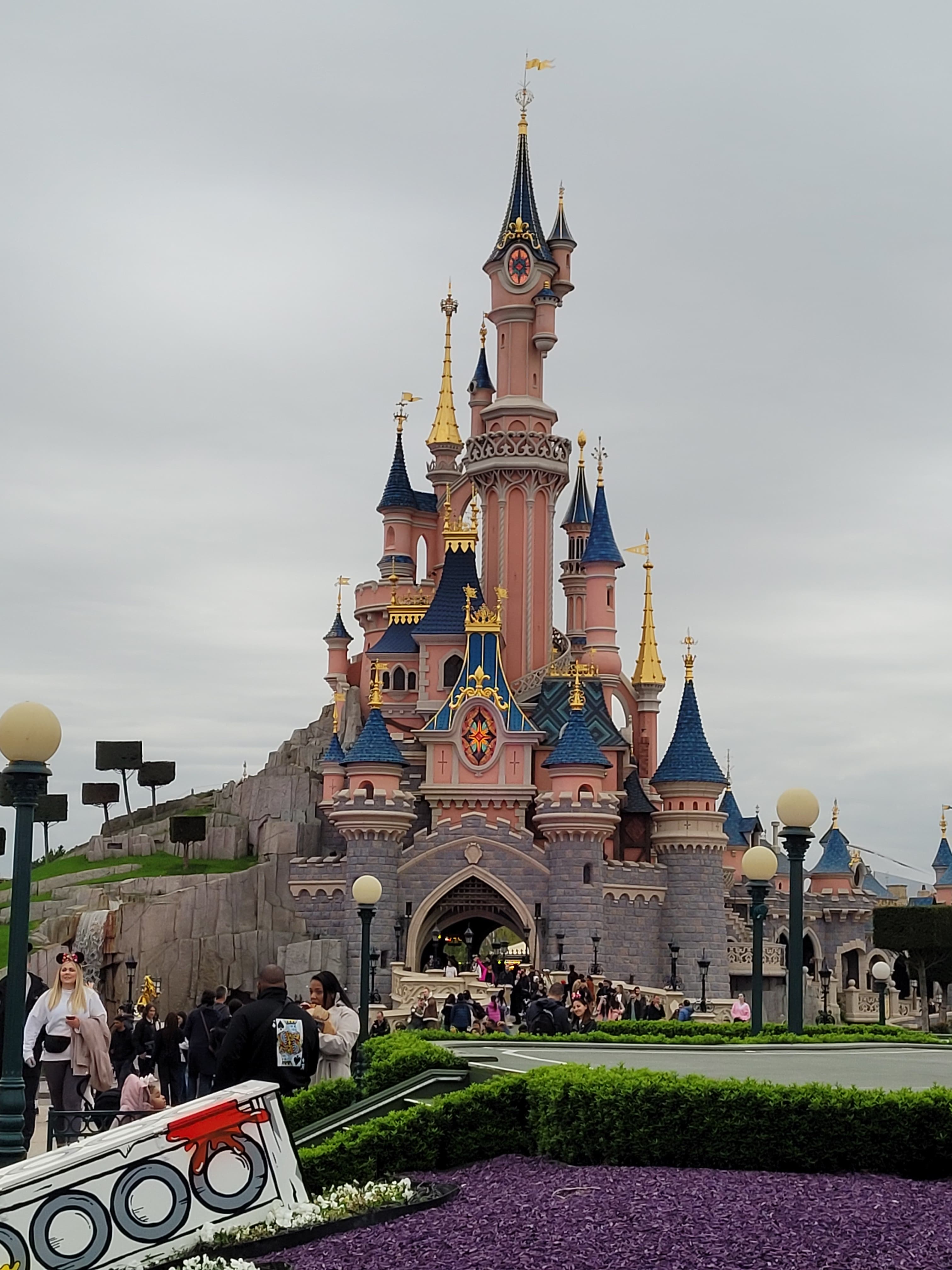 A view of a castle surrounded by landscaping on a sunny day with tourists in the foreground.