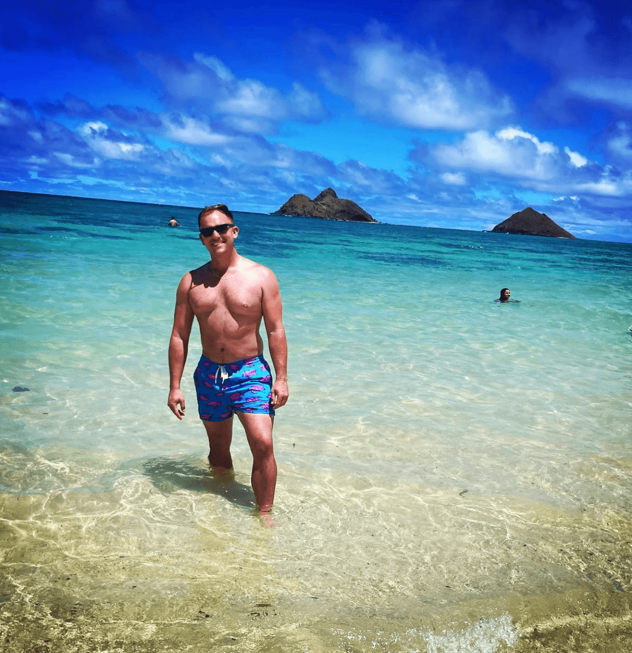 Andrew in swim trunks standing in shallow, clear ocean water on a sunny day