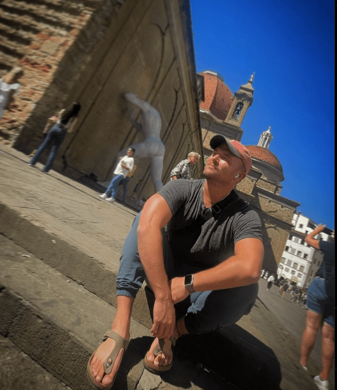 Andrew in a baseball cap and sandals sitting on outdoor steps in front of large monuments on a sunny day