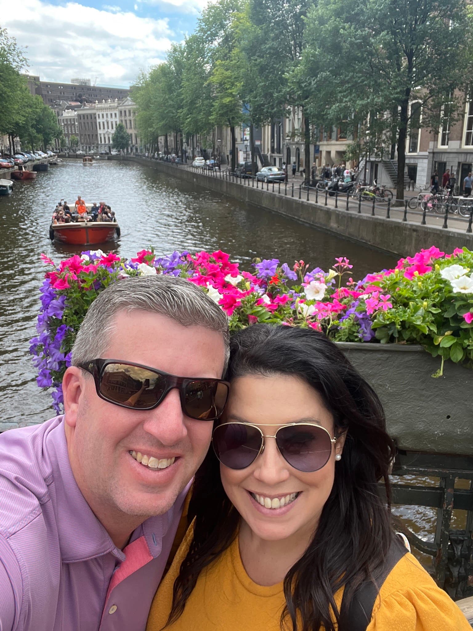 A smiling couple on a boat floating through a canal with pink white and purple flowers behind them.
