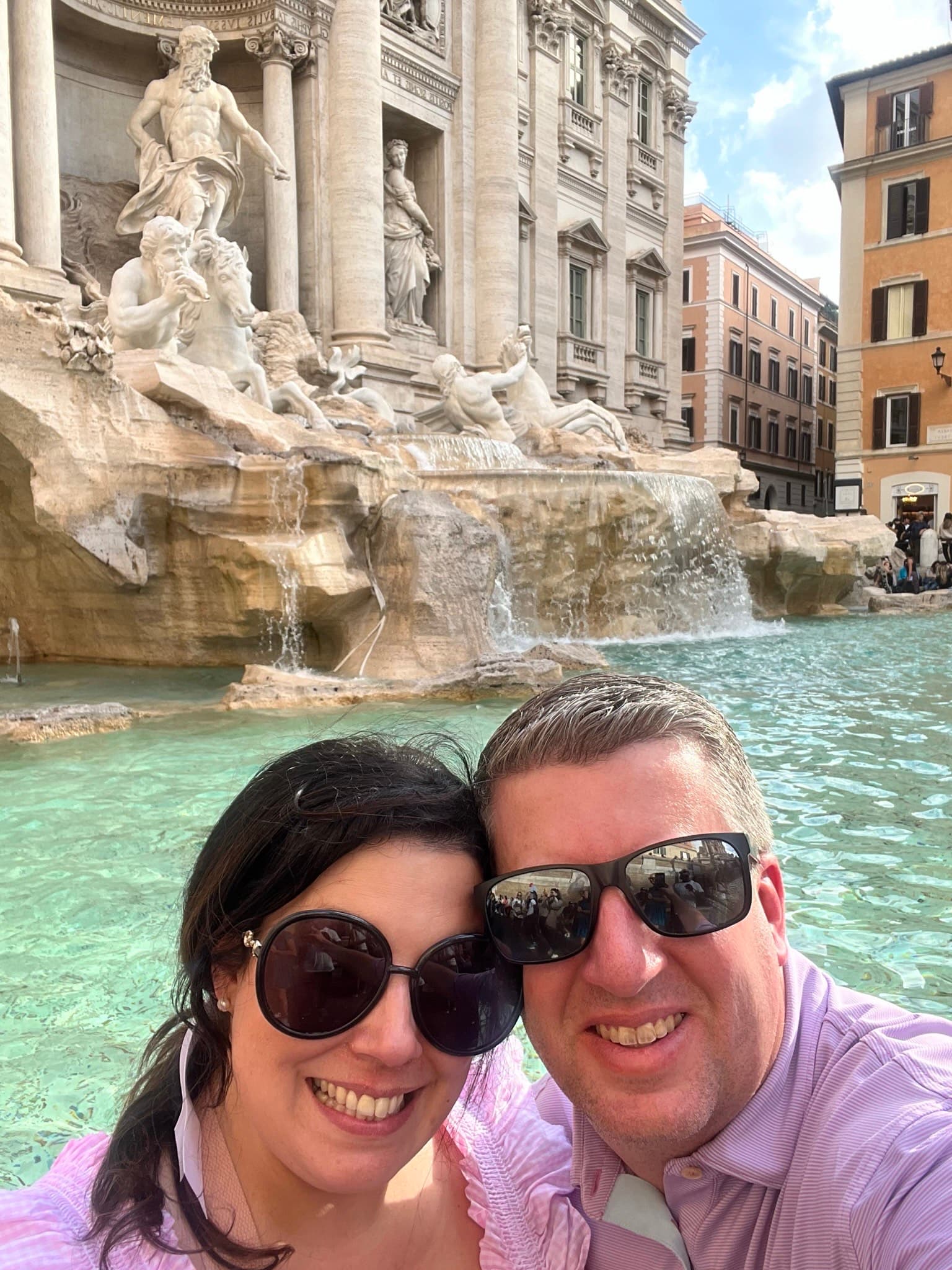 A man and woman smiling in front of a historical stone fountain with carved statues during the day.