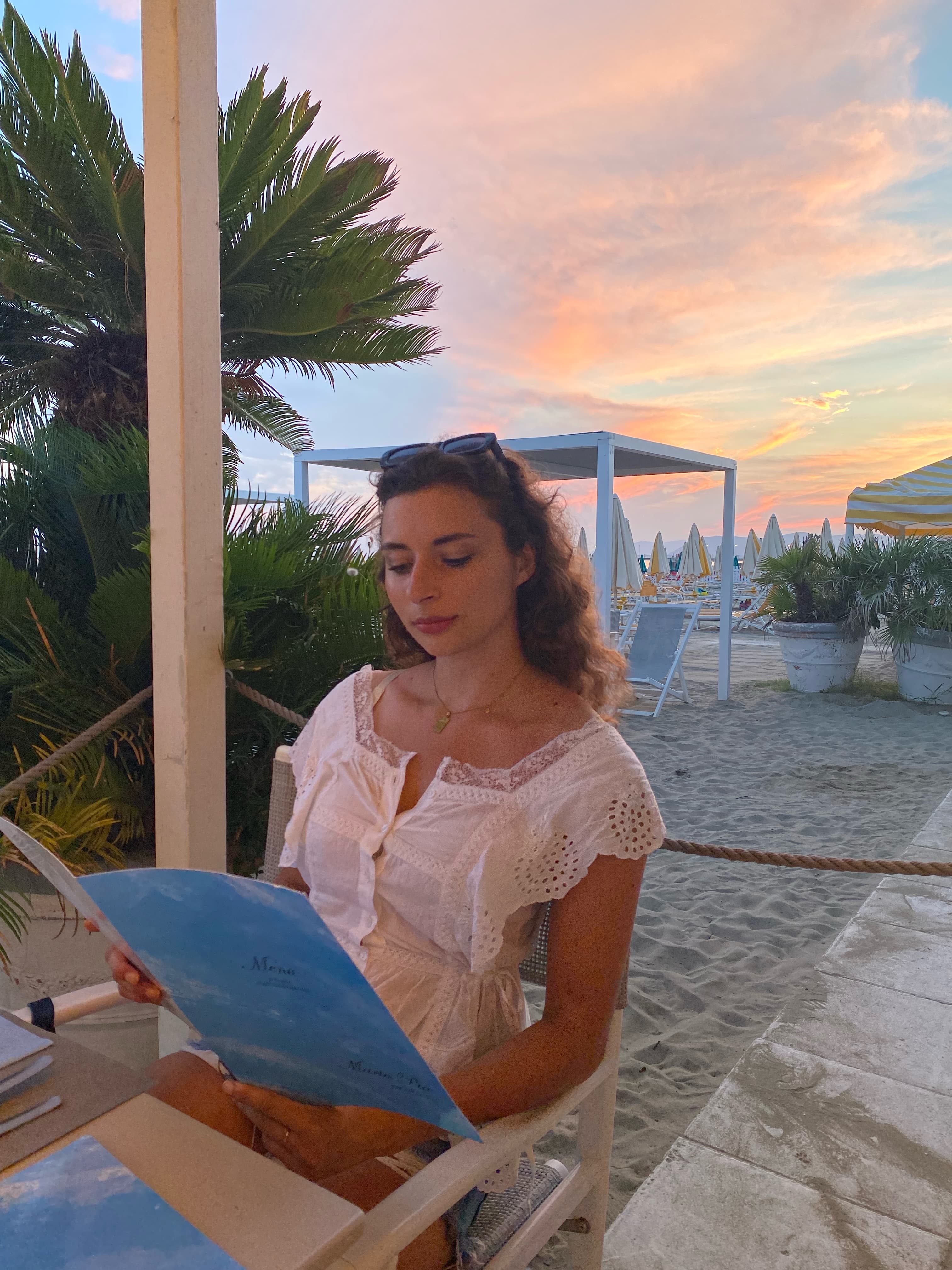 Jordan in a white top reading a menu at a table on the beach with a pastel sunset behind her