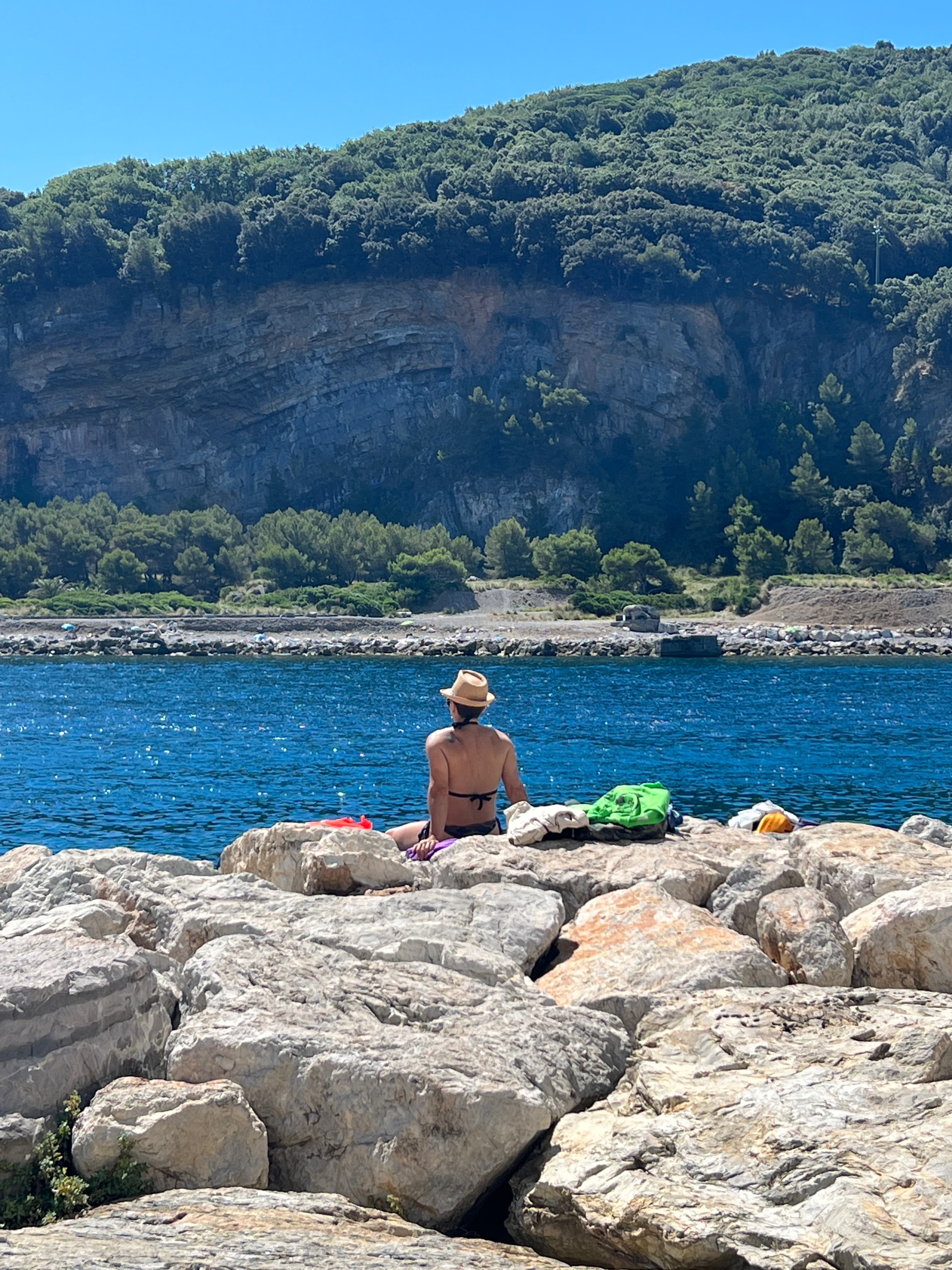 Jordan wearing a hat sitting on rocks facing a calm sea with green cliffs in the distance