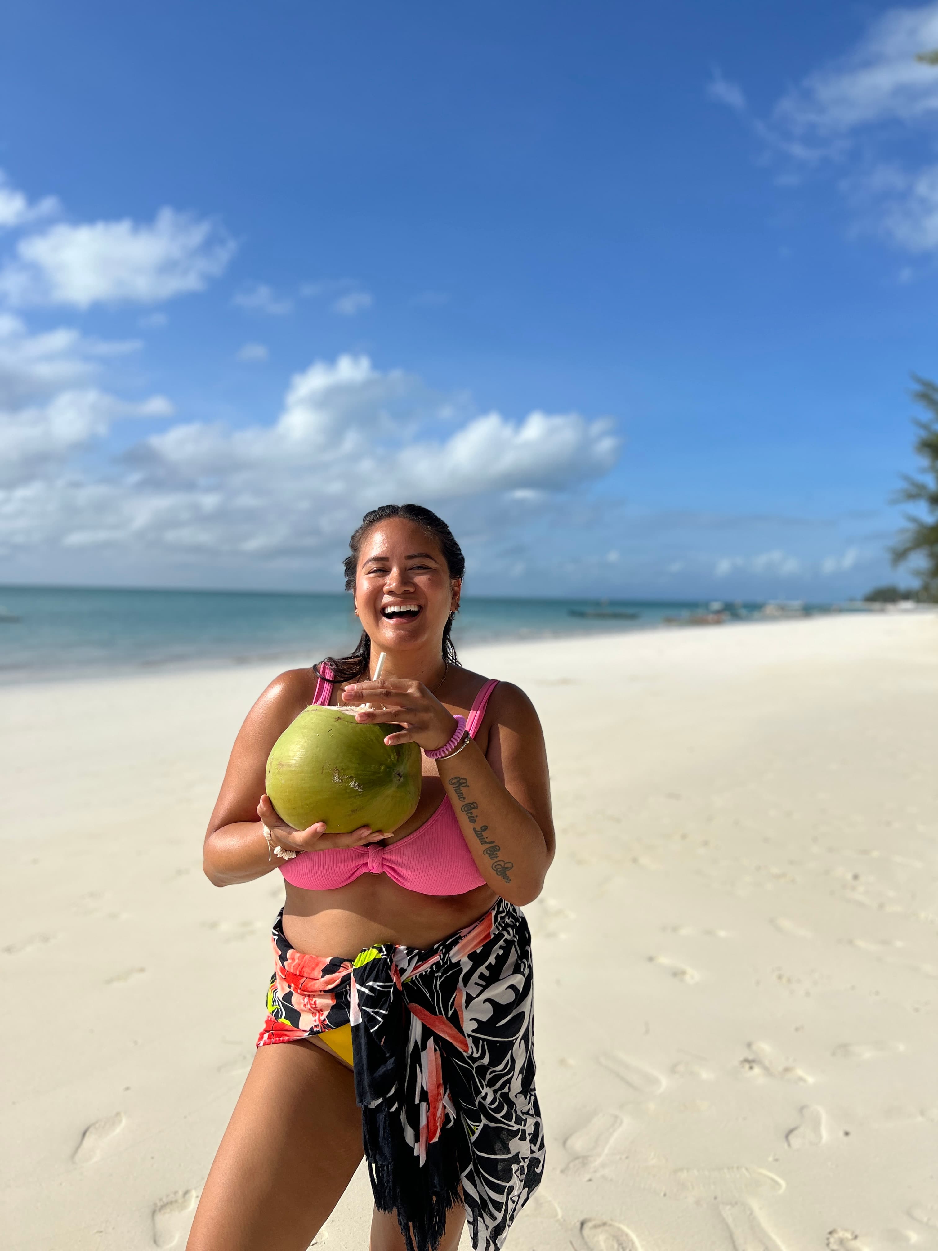 Leah in a bathing suit holding a large coconut on a white sand beach under sunny skies