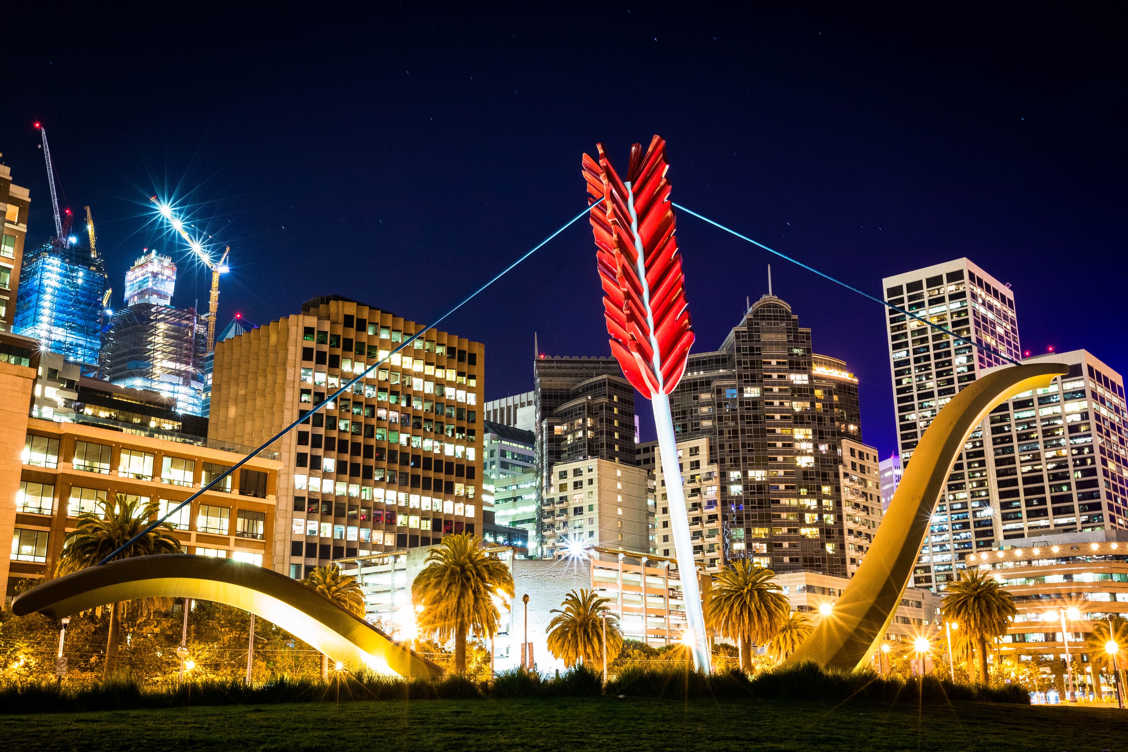 View of a brightly lit city skyline at night with a large structure resembling a bow and arrow in front