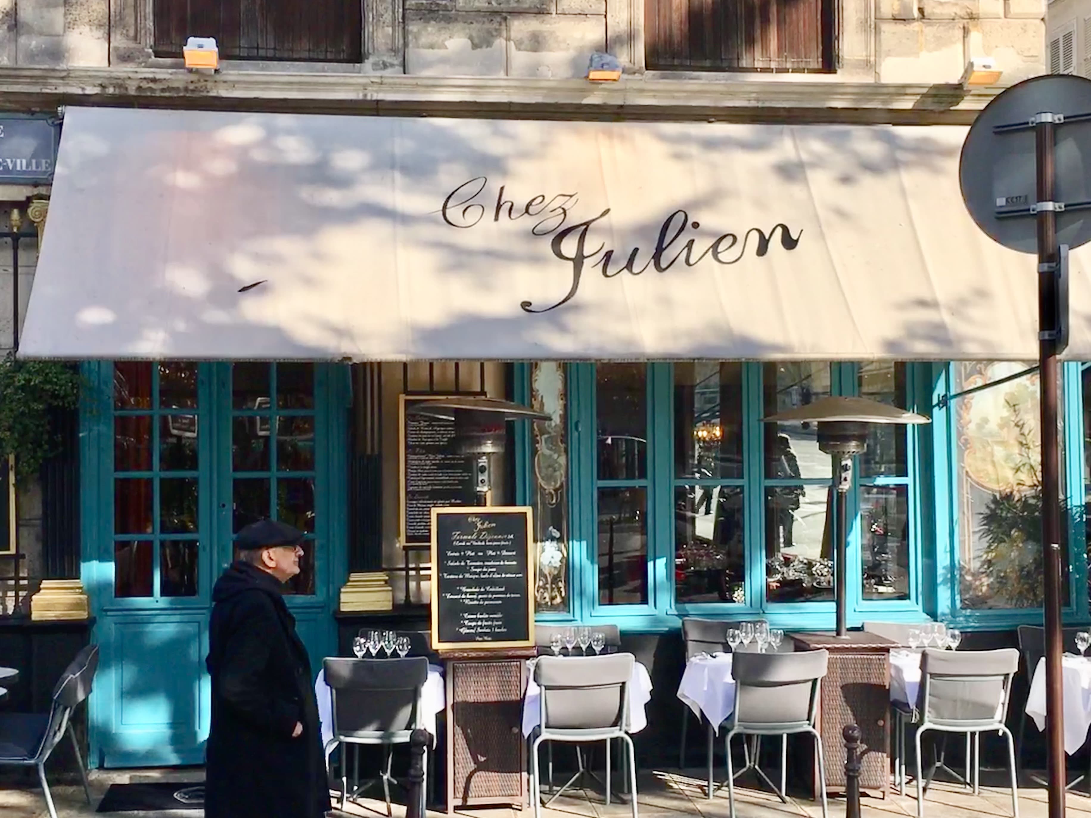 View of a white restaurant awning reading “Chez Julien” hanging over outdoor tables and chairs on a sunny day