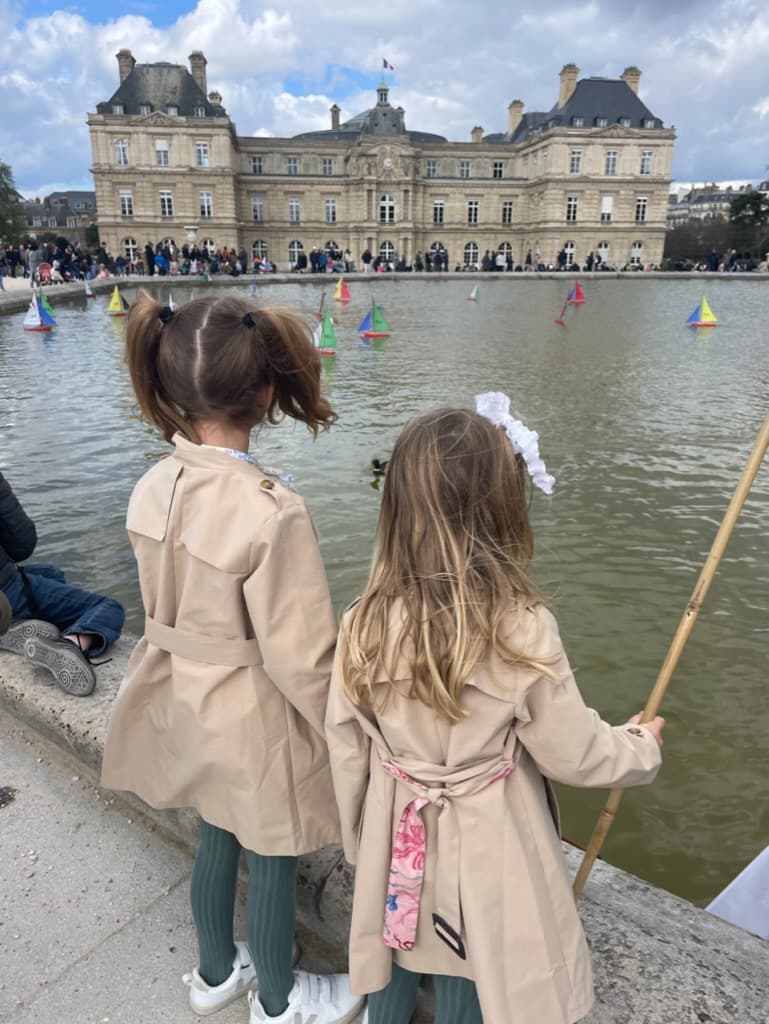 Photo of two girls in beige coats standing in front of a lake during the daytime.