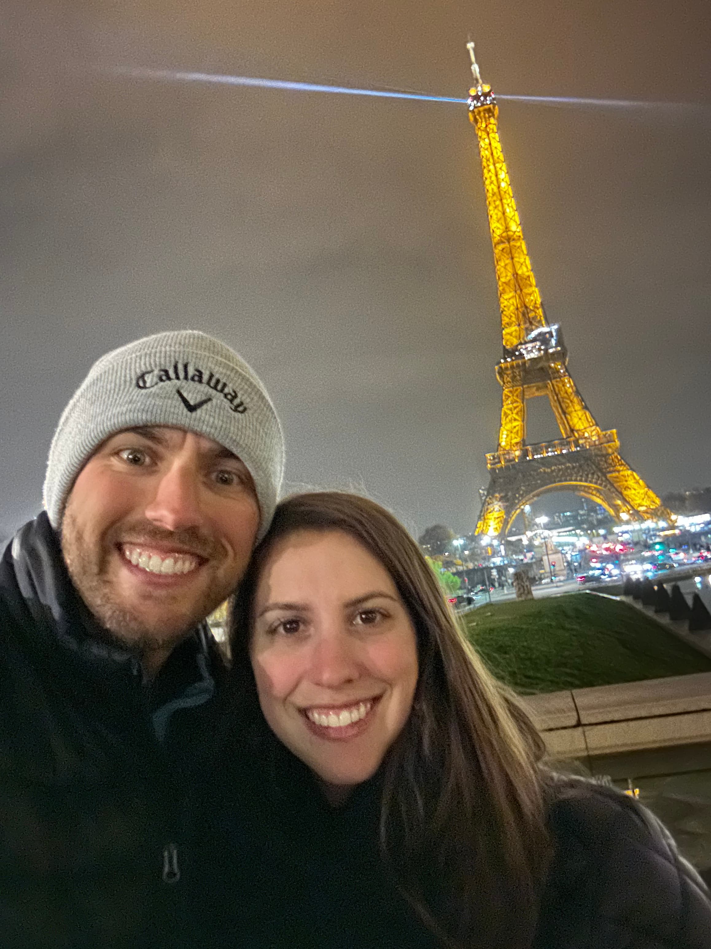 A couple poses for a selfie in front of the lit-up Eiffel Tower at night.