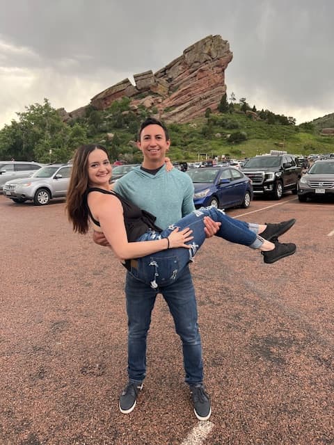 Jaydn’s partner holds her in his arms with a cool rock formation and several cars parked behind them