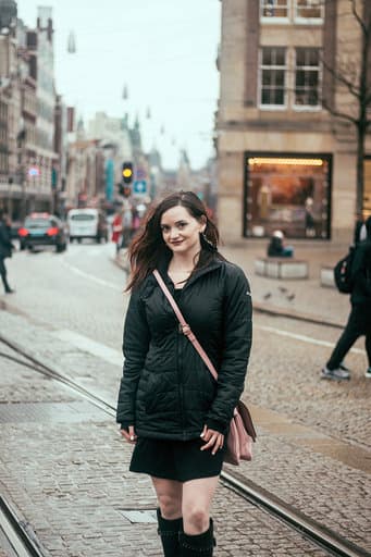 Jadyn in a black coat and boots poses on the street on a cloudy day, pedestrians and cars visible behind her