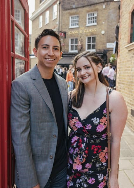 Jadyn in a dress with her arm around her partner in a grey coat posing next to a red telephone box