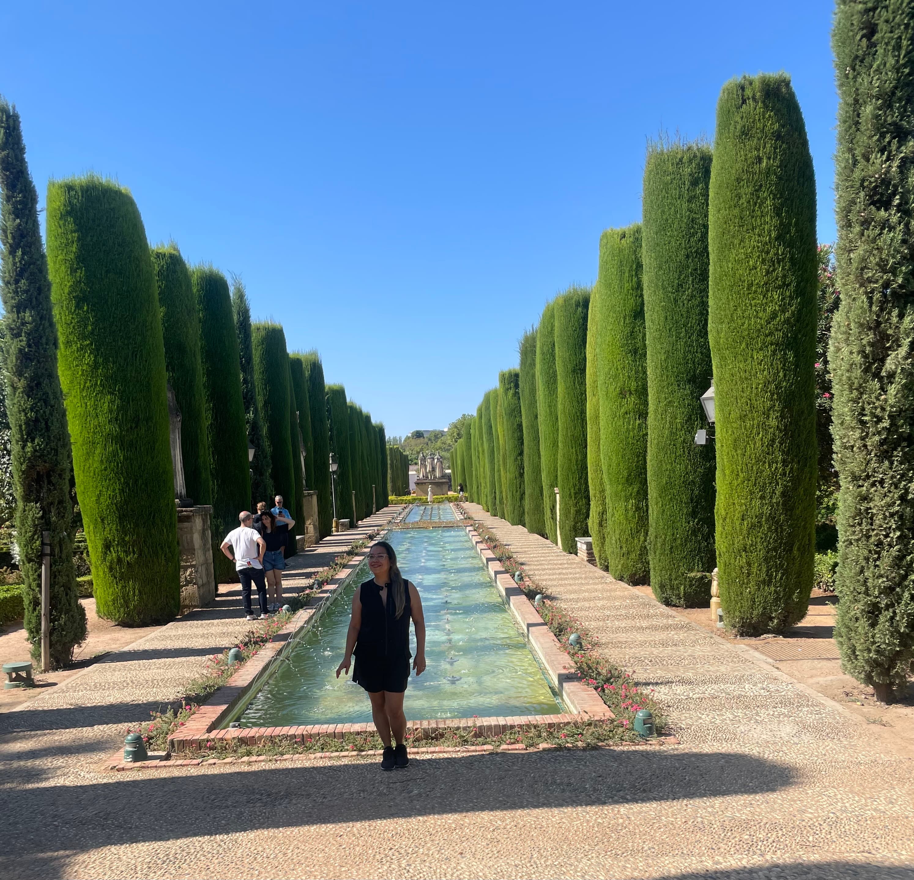 Travel advisor Janeely Mejia standing in a garden with tall, cylinder-shaped bushes in rows down either side of a long pool of water.