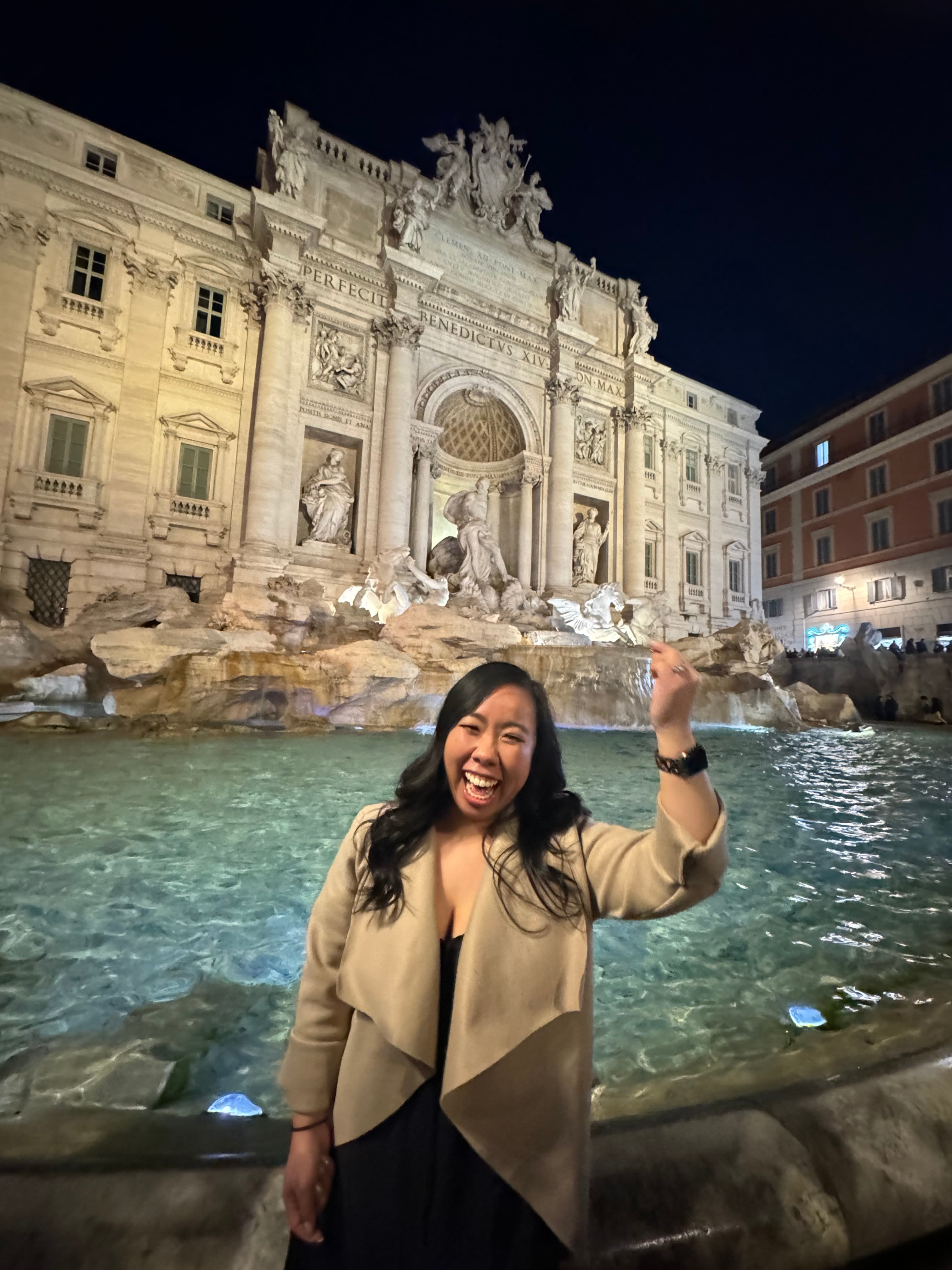 Chelsey in a beige jacket tossing a coin over her shoulder into Rome’s Trevi Fountain at night