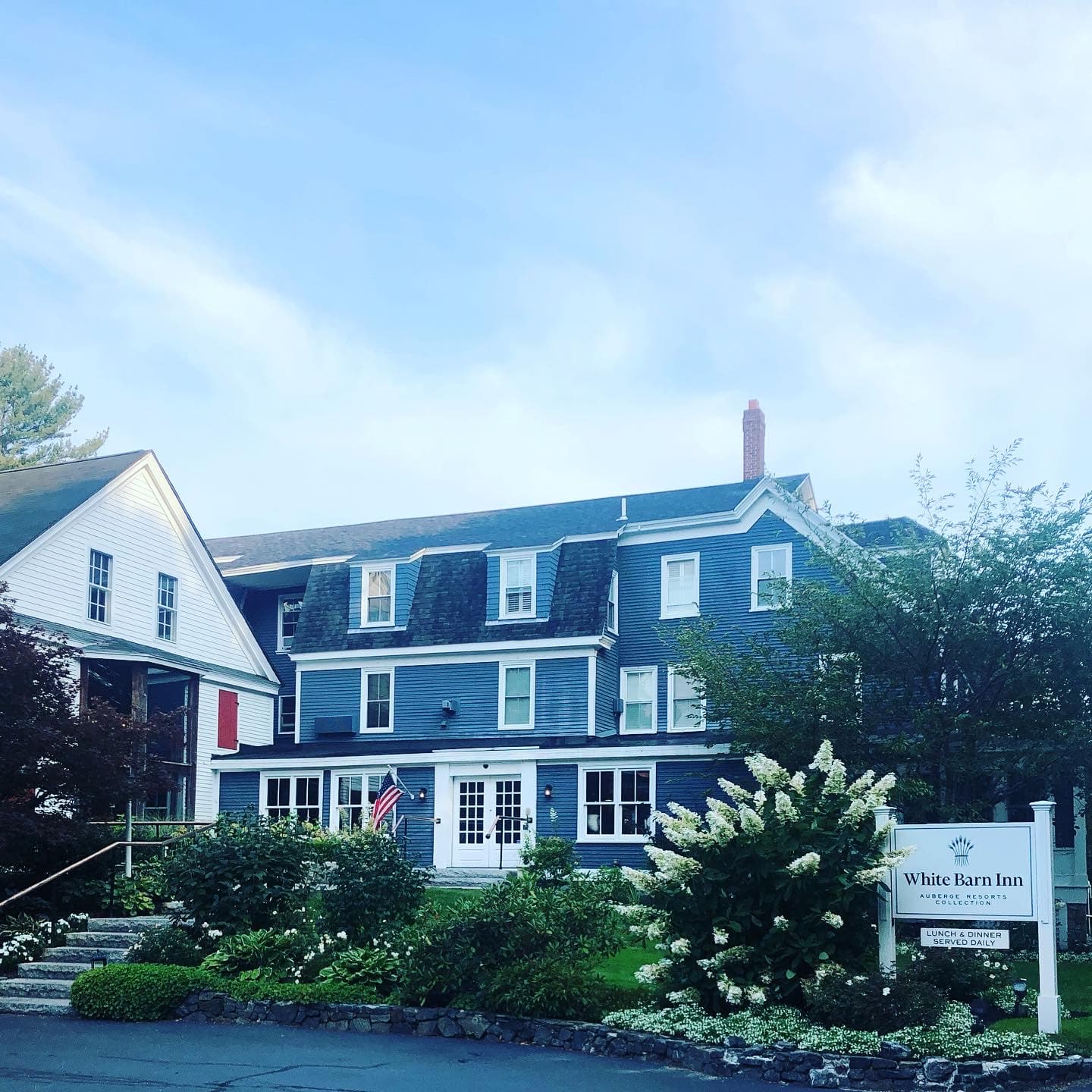 A three story blue building with a chimney and trees and flowers surrounding it during the day.