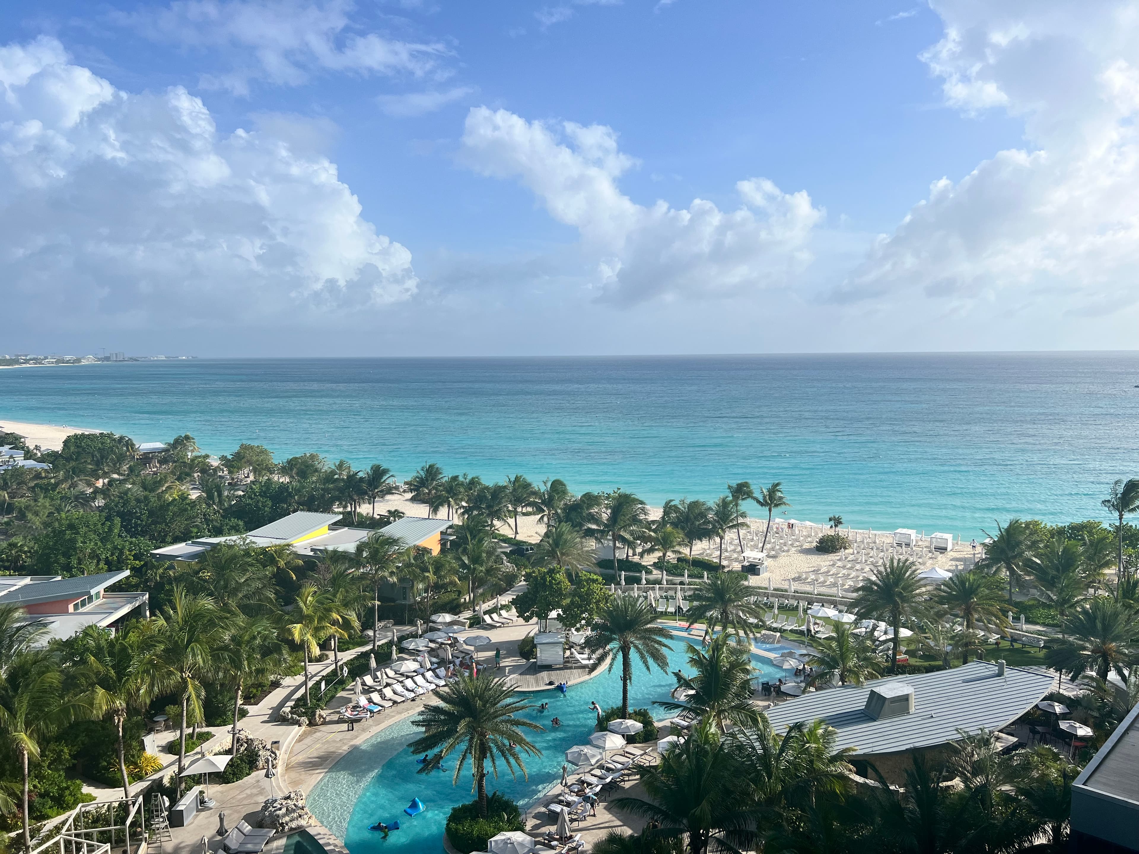 A view overlooking a large resort covered in palm trees, a pool and the ocean during the day.