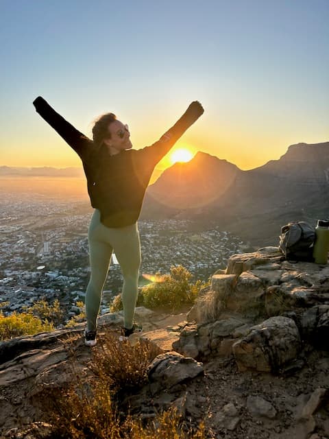 Roberta in a black sweater and arms raised with a beautiful view of a valley and mountains behind her at sunrise