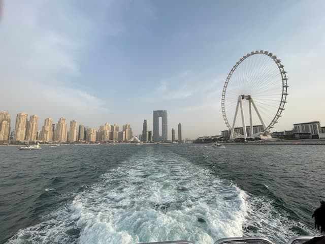 Pretty view of the wake of a boat in a river with a large Ferris wheel to one side and city buildings to the other
