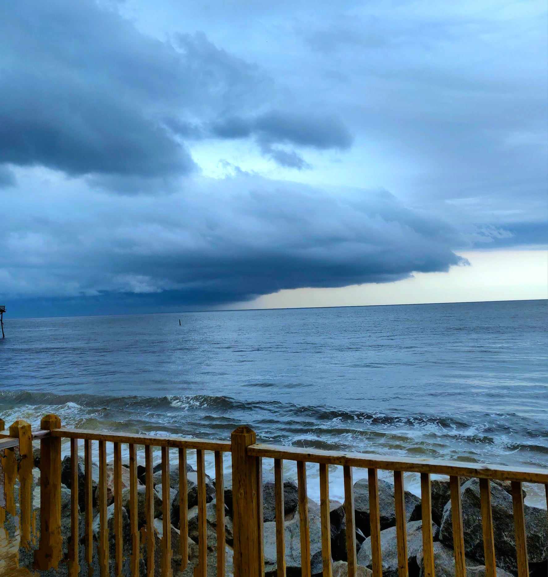 Storm clouds over the ocean with rocks and railing in the foreground.