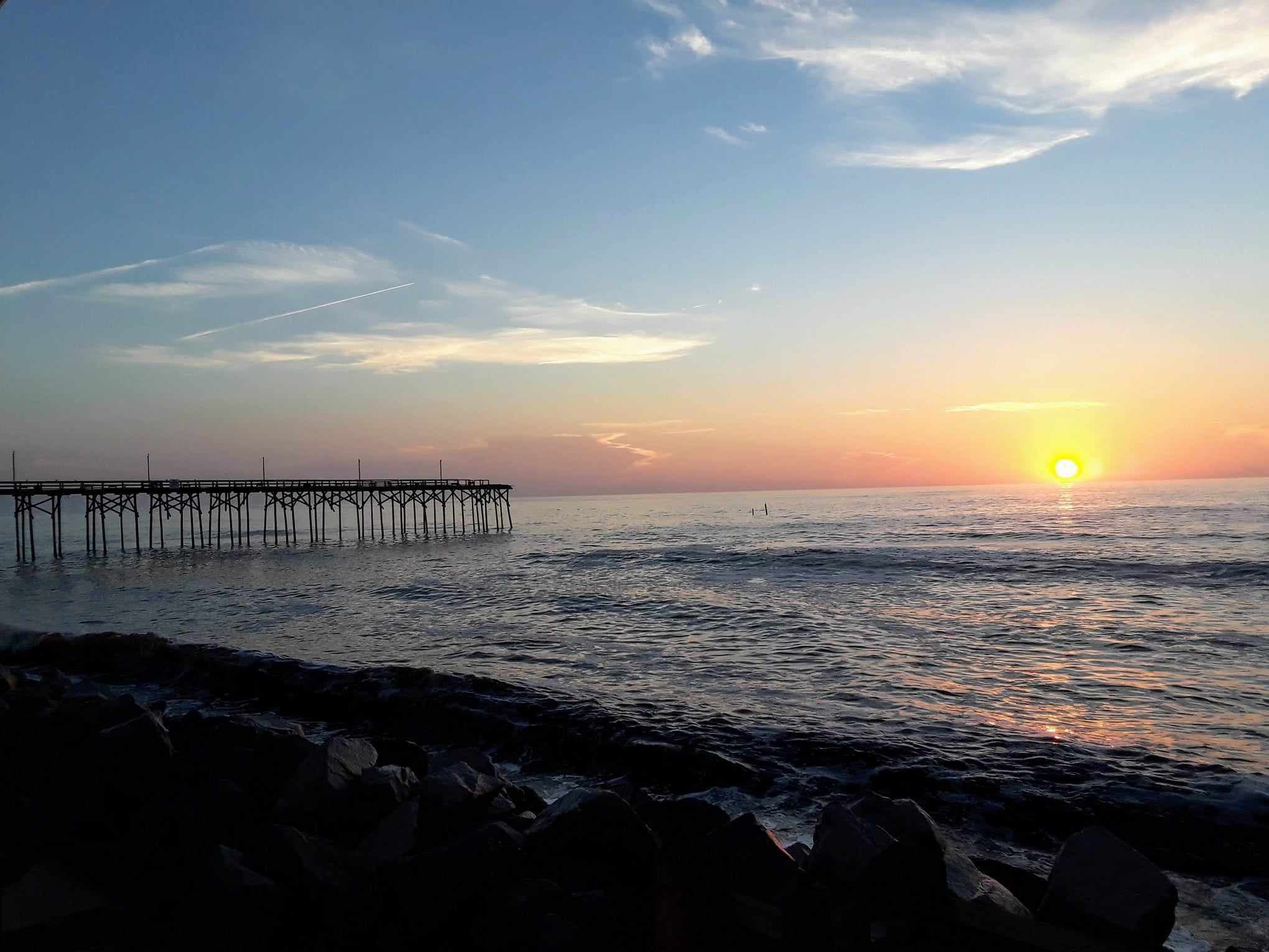 Sun setting over the ocean with beach and a pier in the foreground.