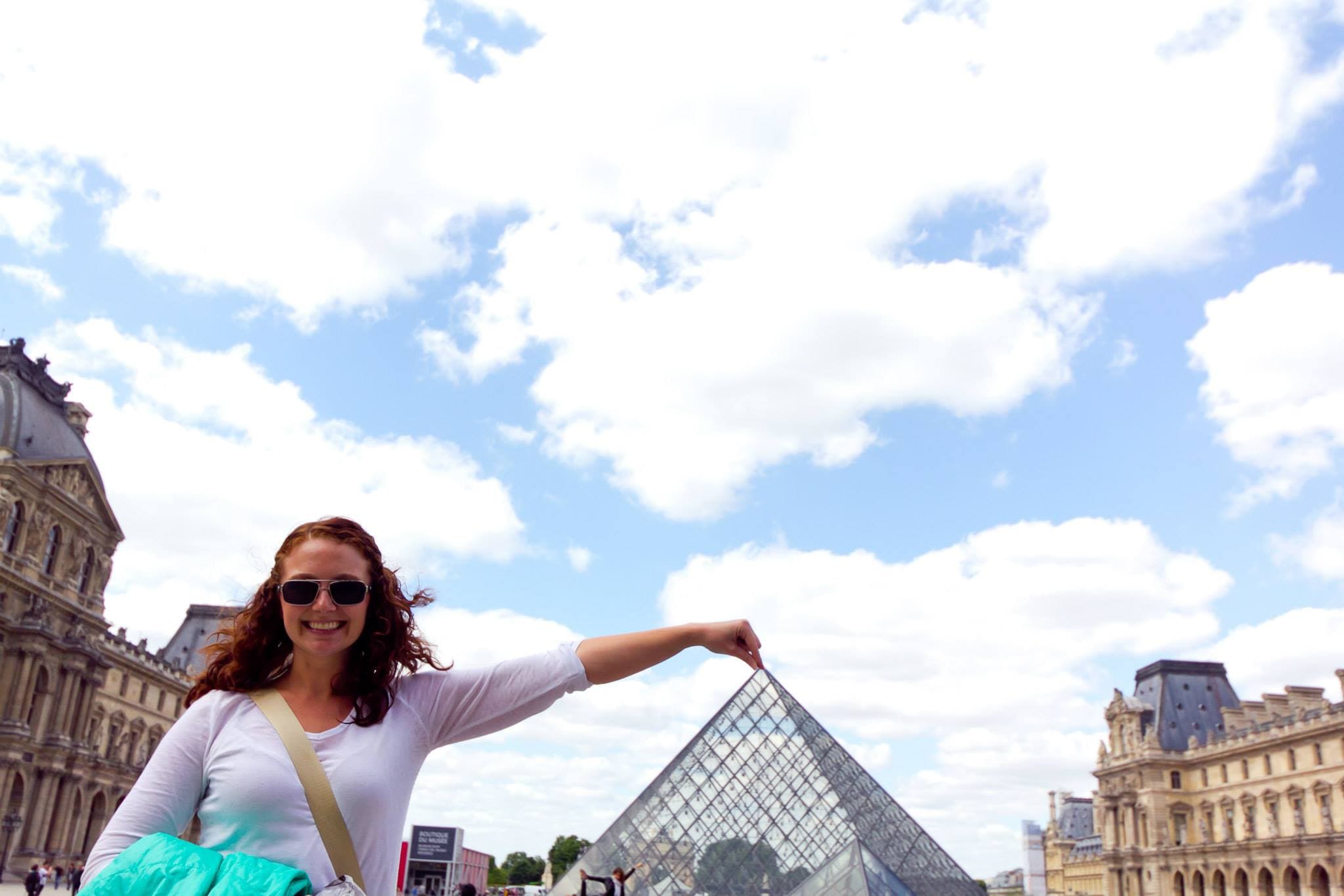 Kayla at the Louvre with her arm outstretched as if she were touching the top of the pyramid