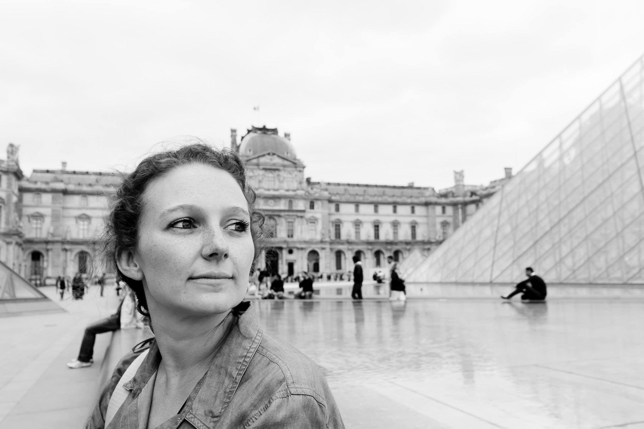 Black and white photo of Kayla in front of the Louvre with the glass pyramid visible behind her