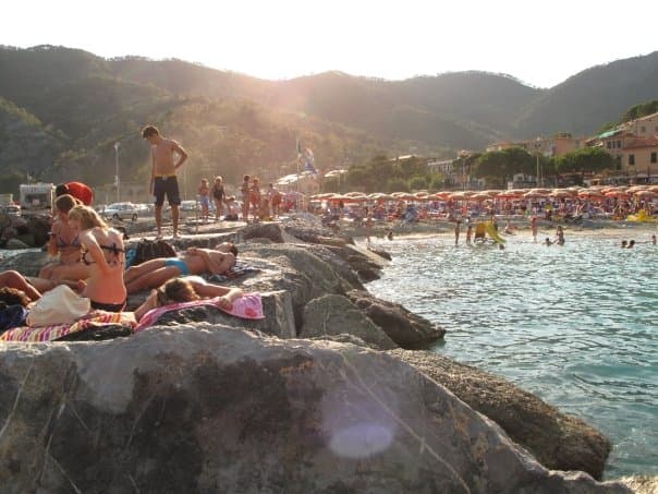 View of people sunbathing on rocks by the sea with mountains behind them at sunset