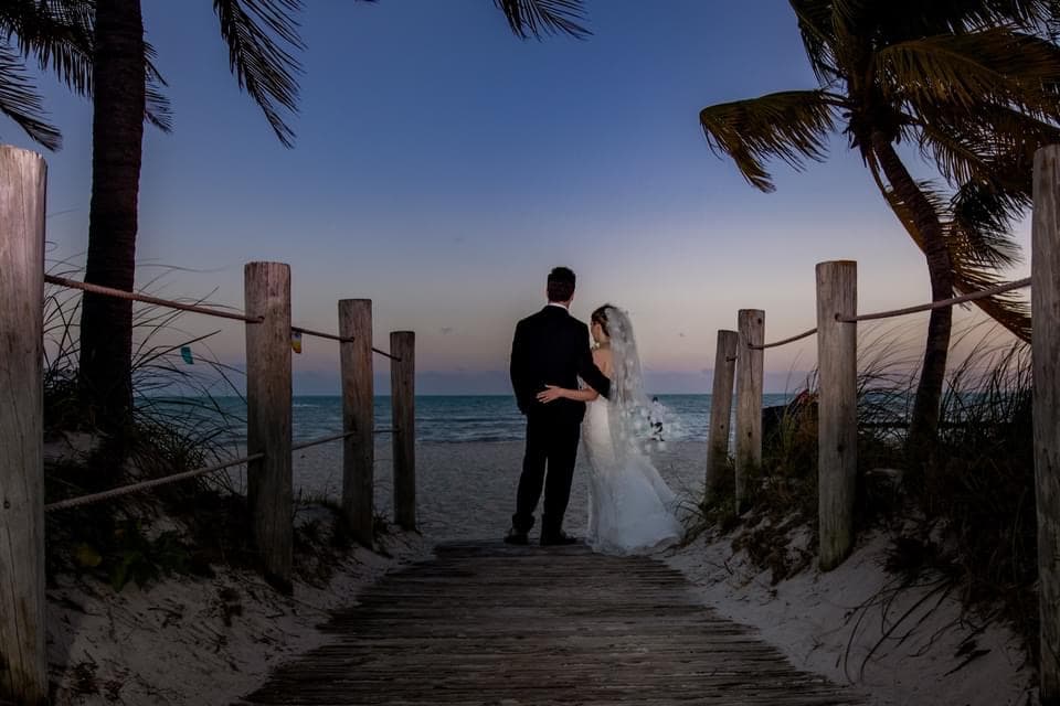 Kimberly in a wedding dress with her arm around her husband standing at the end of a wooden path, the ocean visible behind them