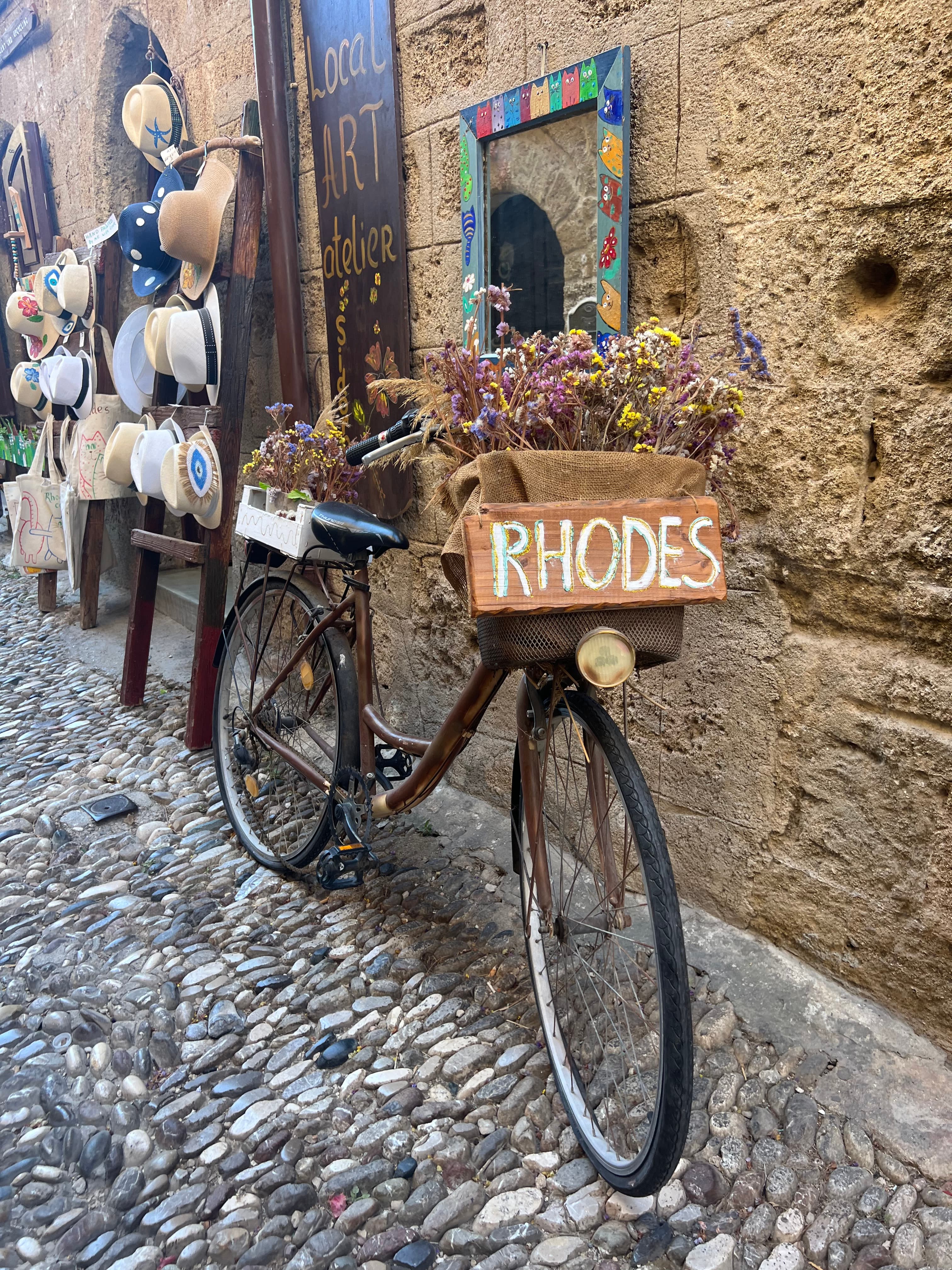 View of a bike with a wooden basket reading ‘RHODES’ and holding flowers against a stone wall