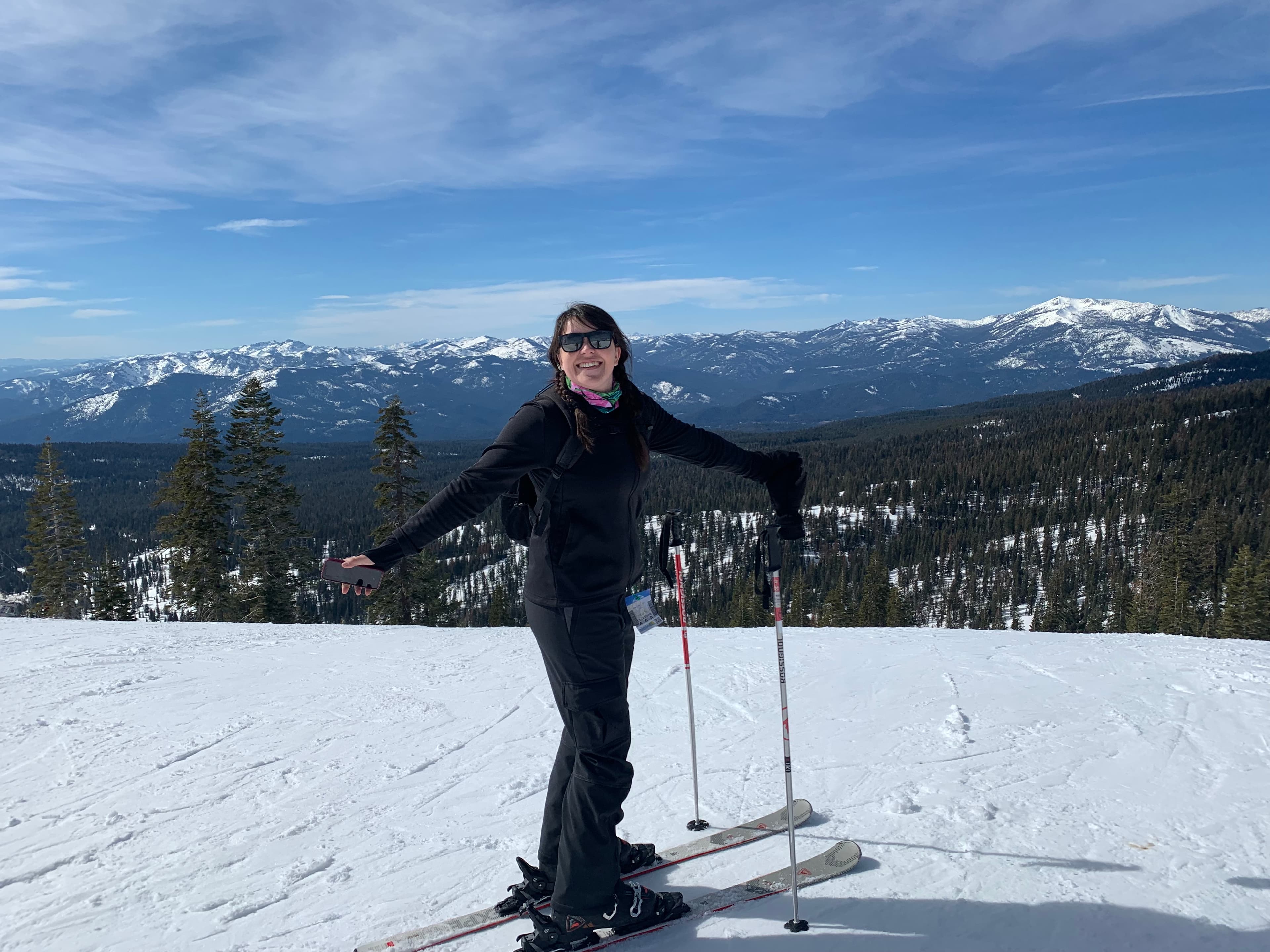 Dani on skis holding poles atop a slope on a sunny day with mountains visible in the distance