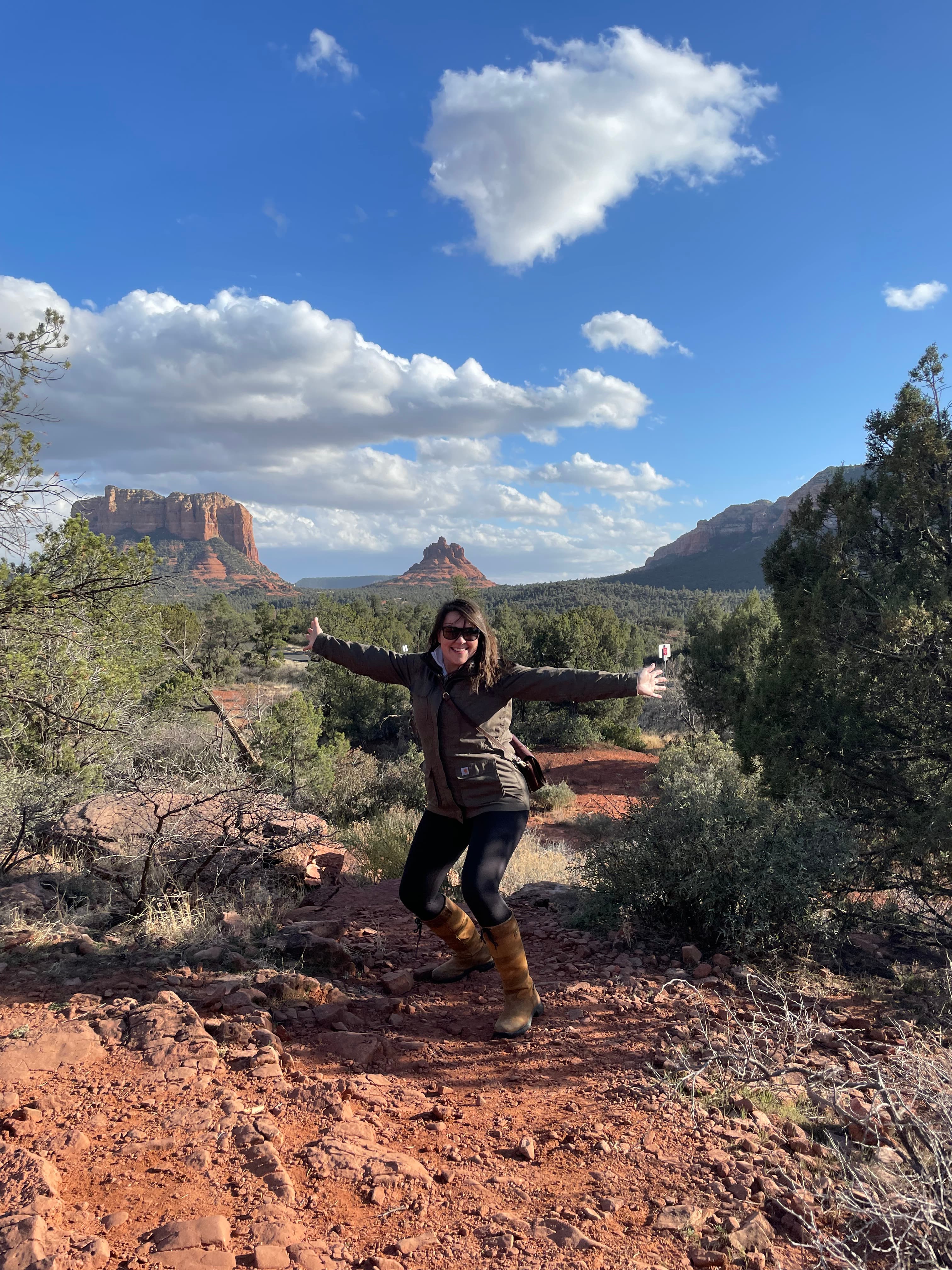 Dani in a black jacket posing with arms spread on a rocky hiking trail under sunny skies