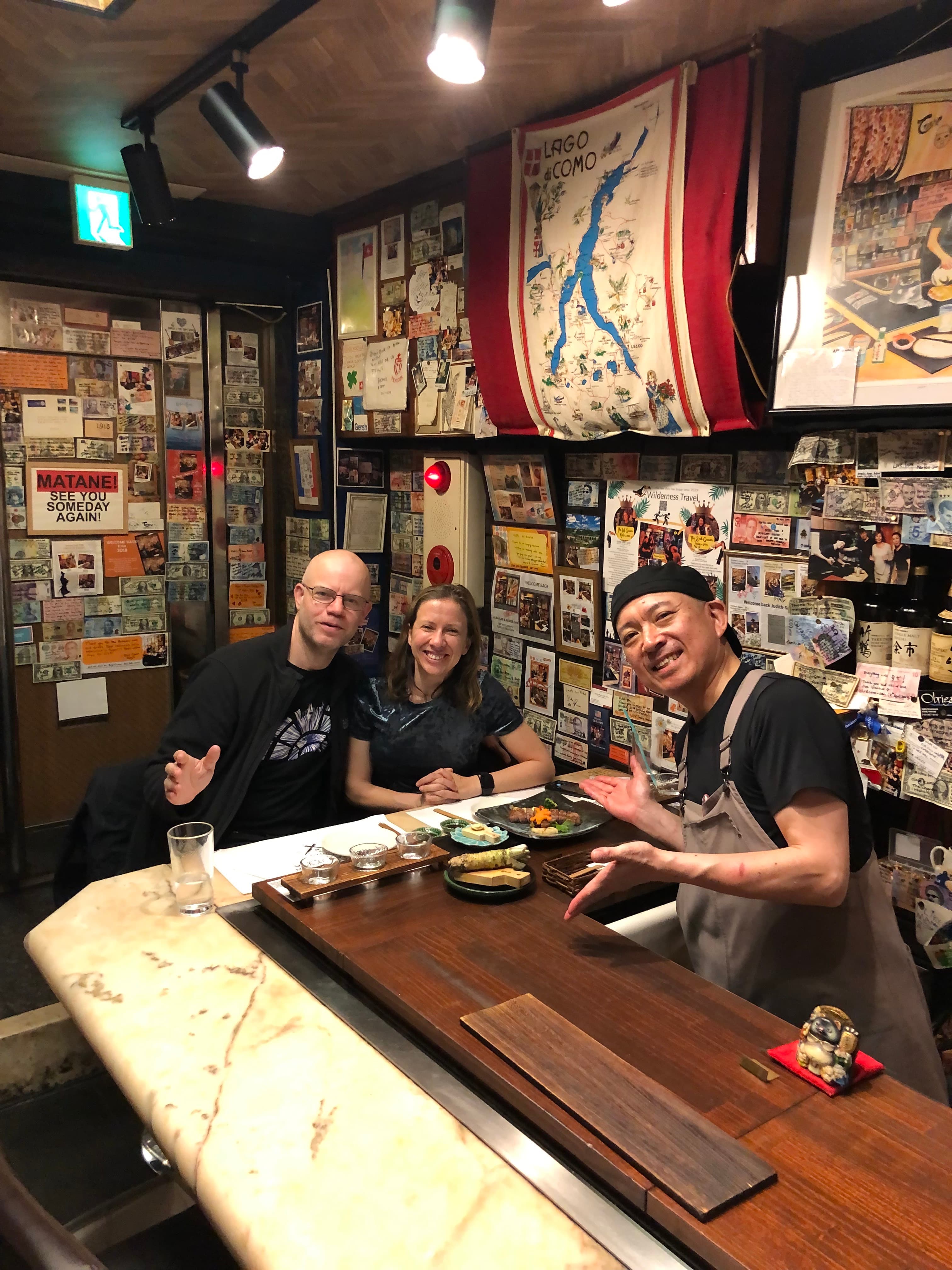 Gerie and a man sitting at a sushi counter, posing with the chef