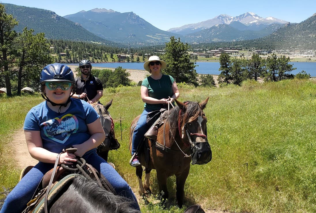 Laura and her family riding horseback through a green field with a beautiful lake and mountain landscape behind them
